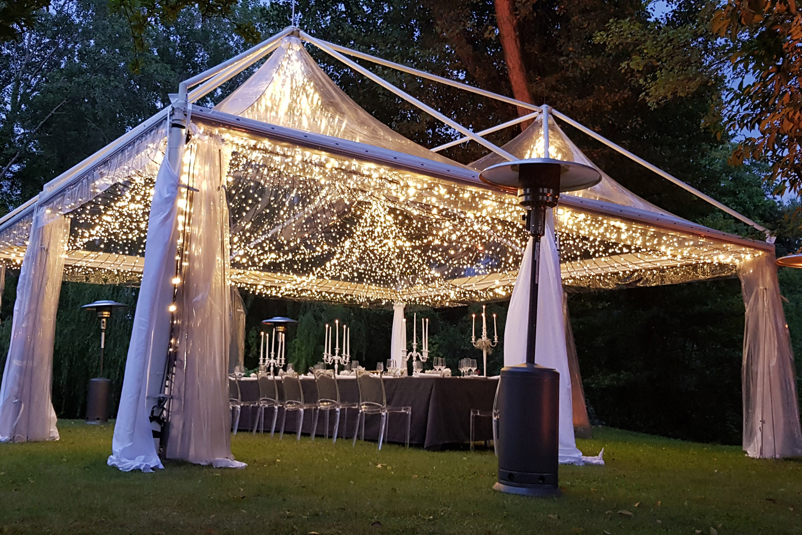 Una tenda esterna trasparente, decorata con lucine, copre un lungo tavolo da pranzo apparecchiato con candele ed eleganti tovaglie. Intorno alla tenda si trovano alte stufe, e la scena è circondata da alberi e prato al tramonto.