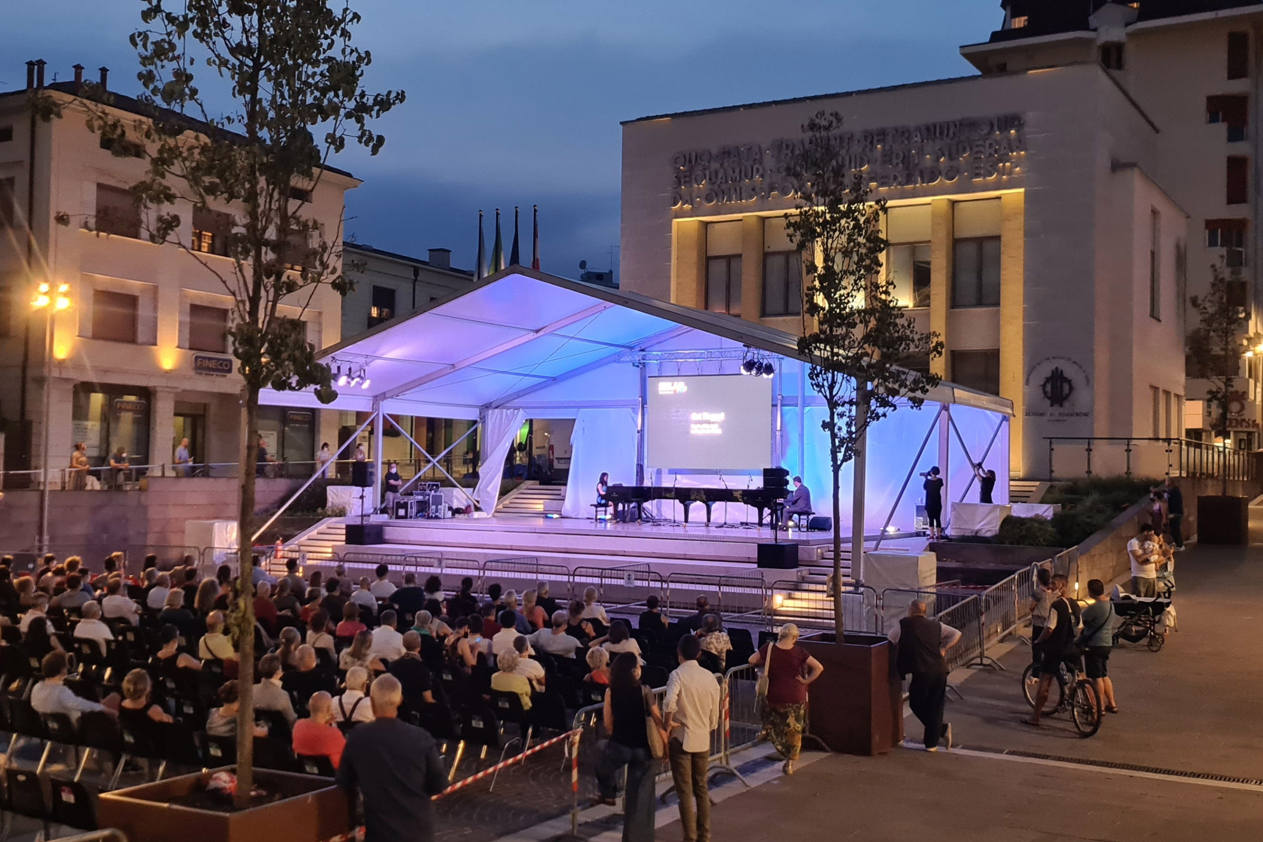 Un concerto serale all'aperto con un pubblico seduto davanti a un palco sotto una tenda bianca. Sul palco ci sono un pianoforte a coda e uno schermo, con un edificio illuminato sullo sfondo. Alberi e luci circondano la piazza.