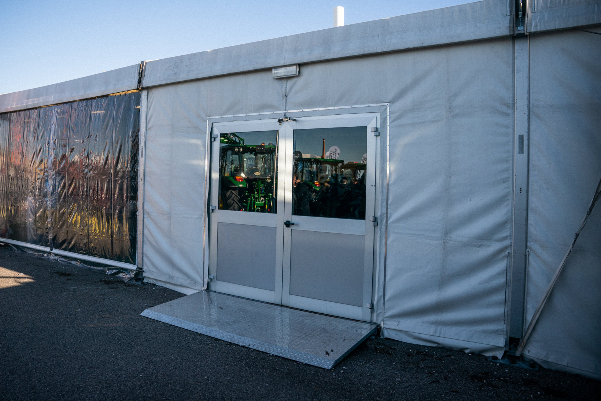 Una grande tenda bianca con doppie porte in vetro e una rampa di metallo è adagiata su una superficie pavimentata; all'interno, i riflessi mostrano trattori o macchinari attraverso le porte.