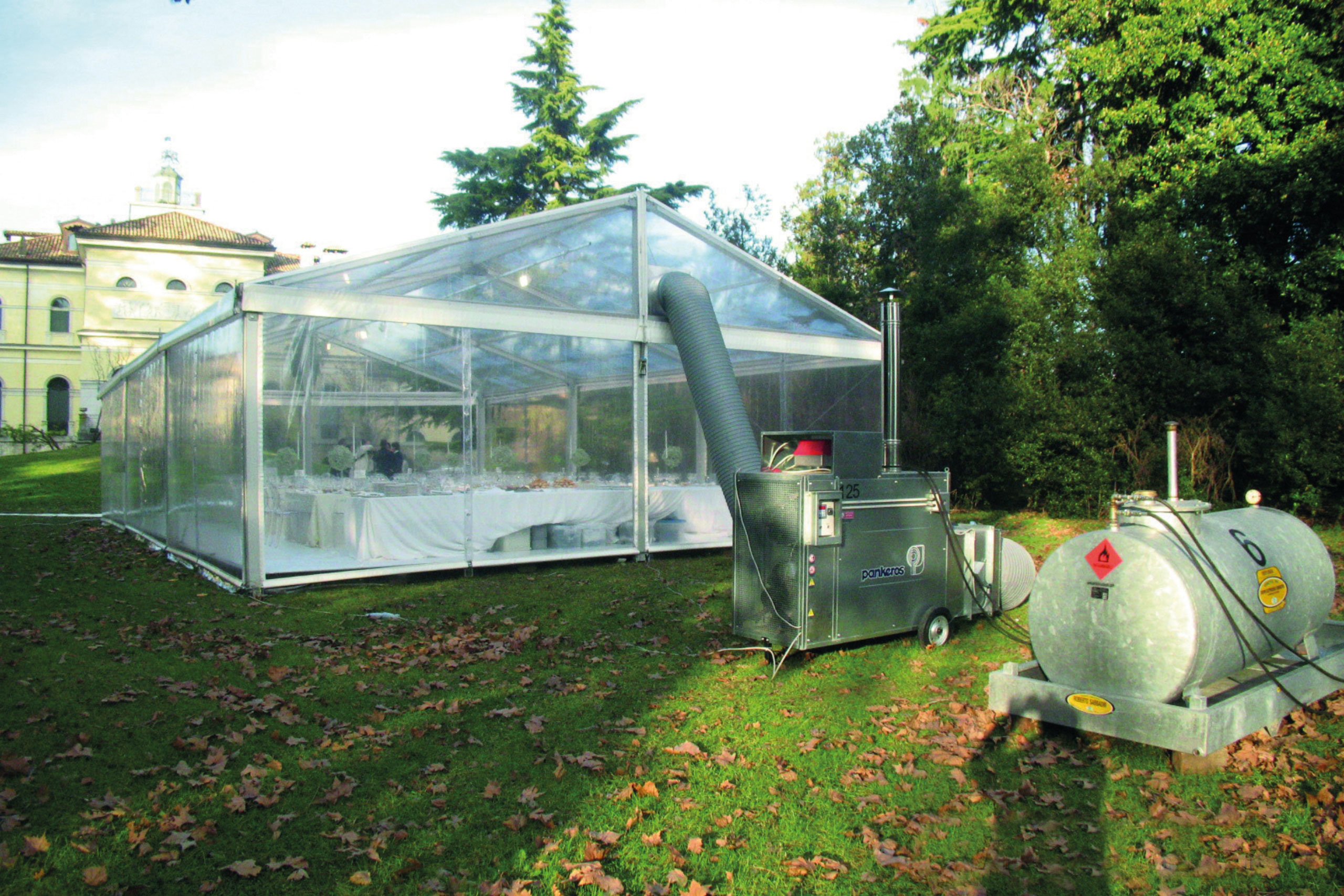 Una grande tenda trasparente è allestita su un'area erbosa, con tavoli e sedie all'interno per l'evento. Nelle vicinanze, un grande generatore metallico e un serbatoio sono posizionati a terra, con un tubo che collega il generatore alla tenda.