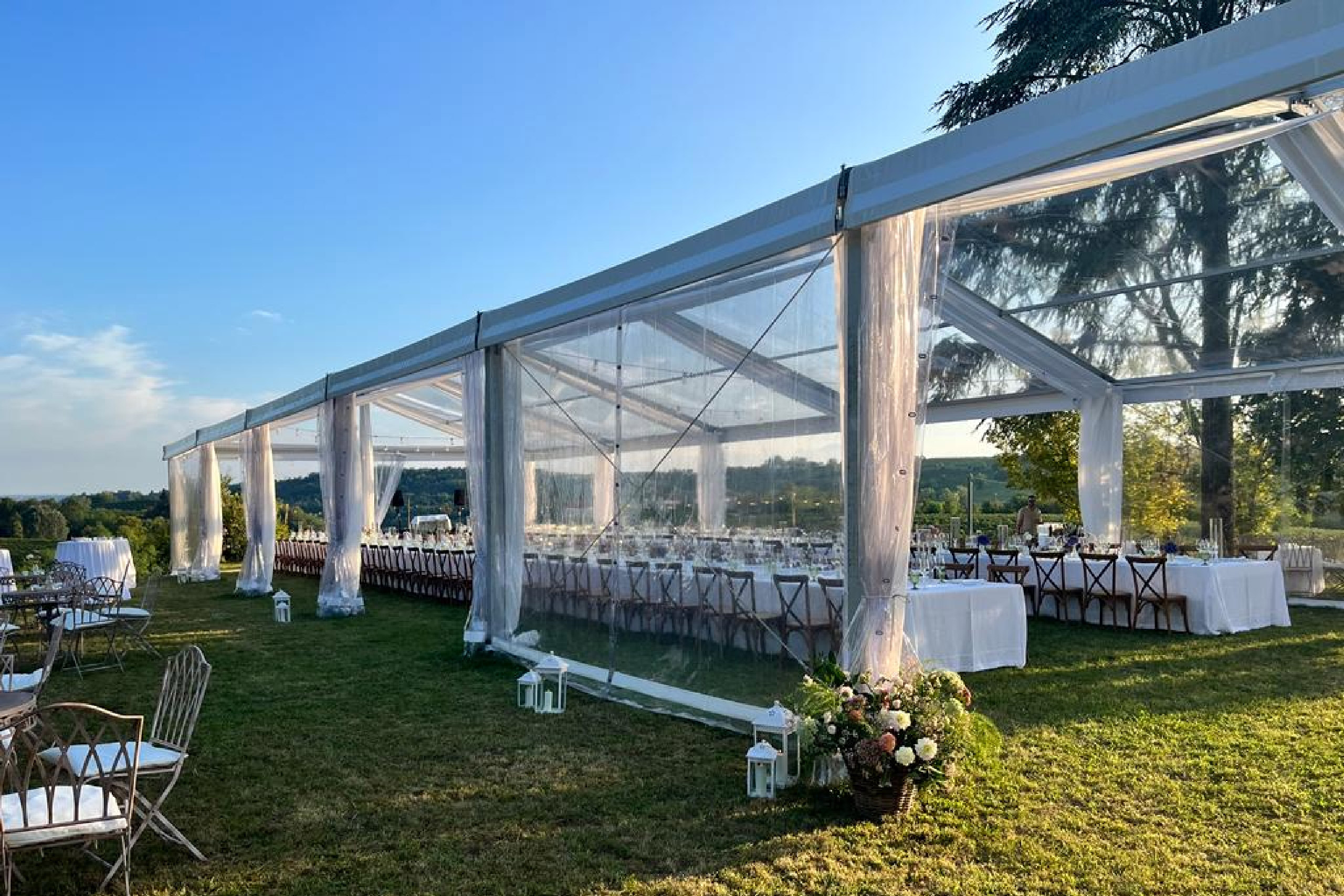 Una tenda trasparente allestita all'aperto sull'erba con lunghi tavoli da pranzo, tovaglie bianche e sedie di legno, decorata con fiori e lanterne, che si affaccia su un paesaggio suggestivo sotto un cielo azzurro.