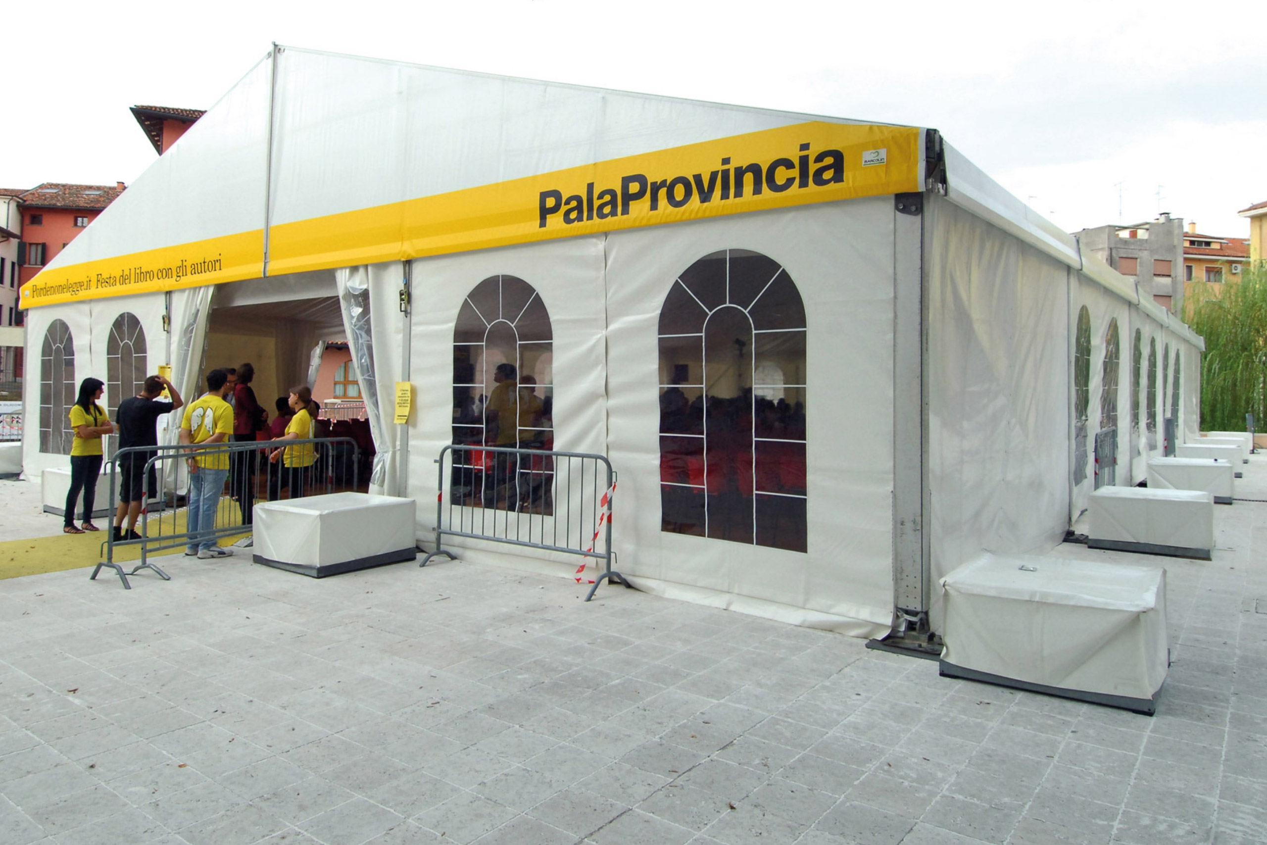 Un grande tendone bianco per eventi con la scritta "PalaProvincia" e persone radunate vicino all'ingresso, alcune delle quali indossano magliette gialle. Barriere metalliche e panchine coperte sono posizionate all'esterno, sull'area pavimentata.