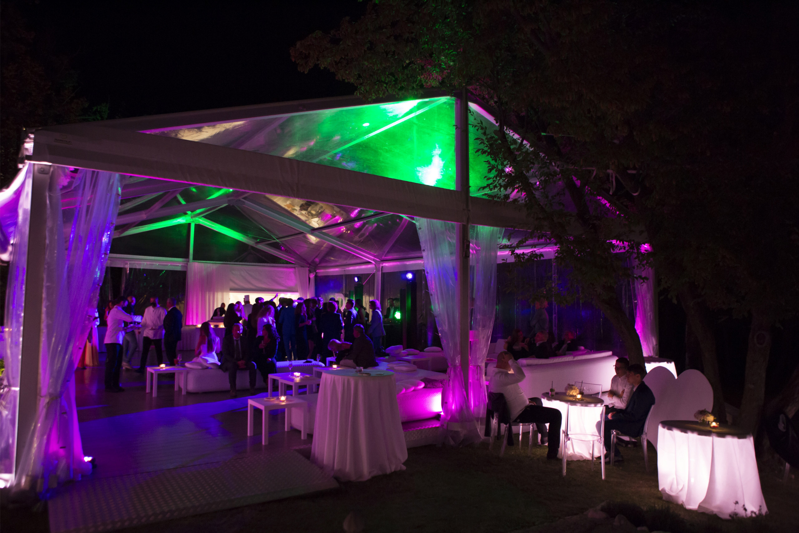 Una festa notturna all'aperto sotto una tenda trasparente con luci colorate viola e verdi. Gli ospiti si mescolano al centro, mentre altri siedono su tavoli e sedie bianche lungo i bordi della tenda. Gli alberi incorniciano la scena festosa.