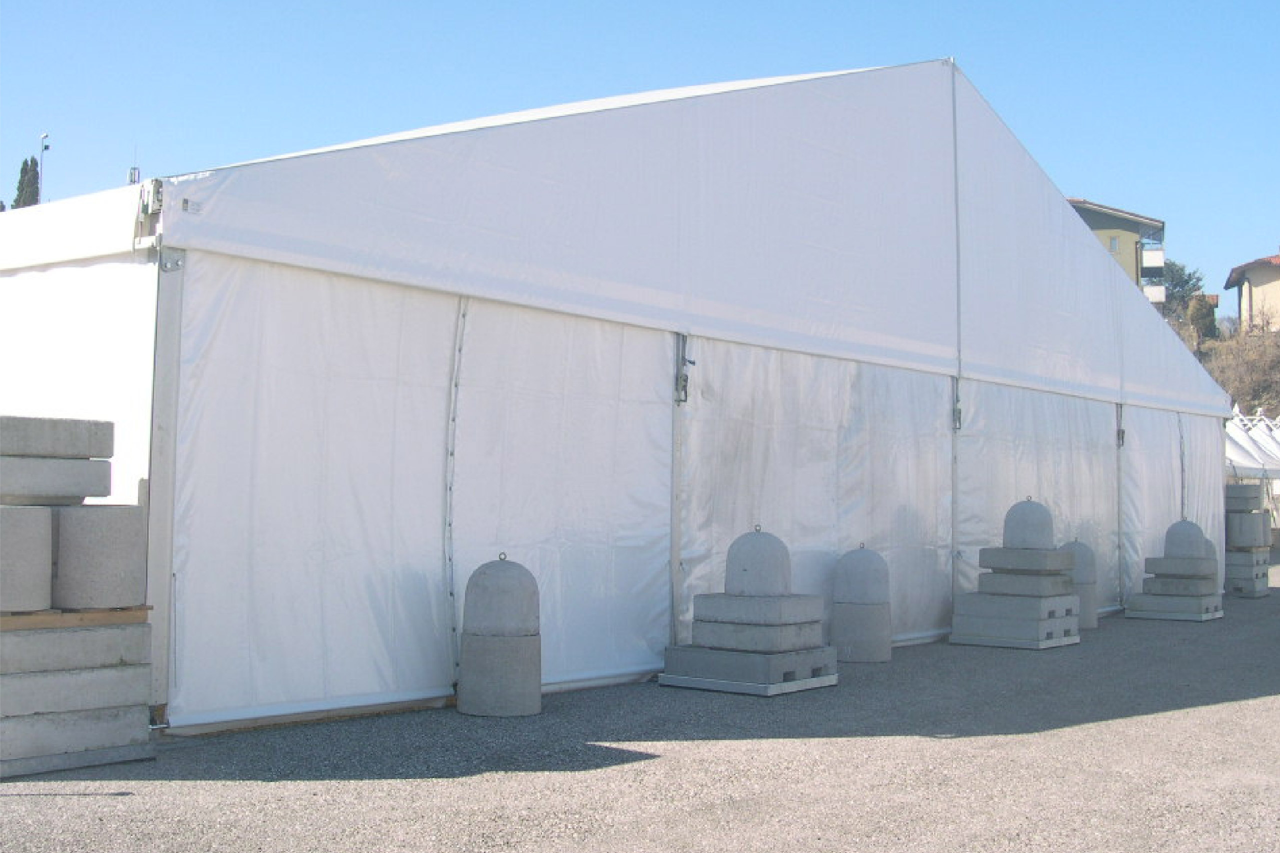 Una grande tenda temporanea bianca con blocchi di cemento appesantiti alla base, installata all'aperto su una superficie di ghiaia sotto un cielo azzurro e terso.