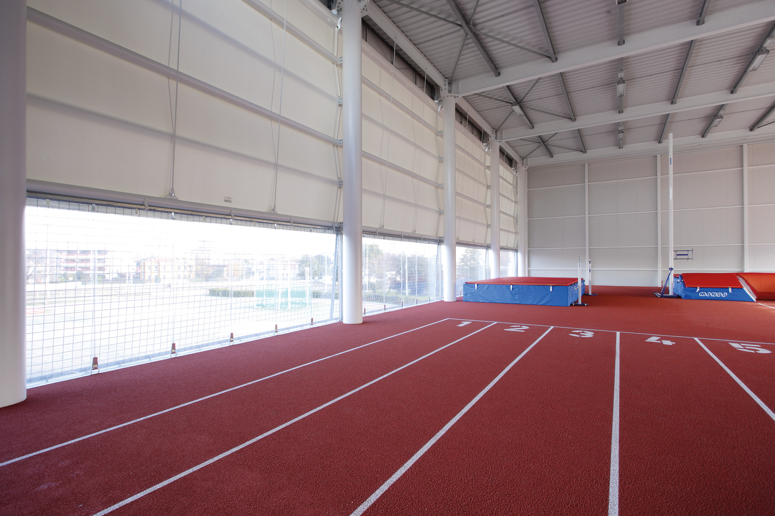 Pista di atletica al coperto con corsie rosse numerate da 1 a 5, grandi finestre su un lato che lasciano entrare la luce naturale e materassini per il salto con l'asta visibili alla fine della pista, vicino a un muro bianco.