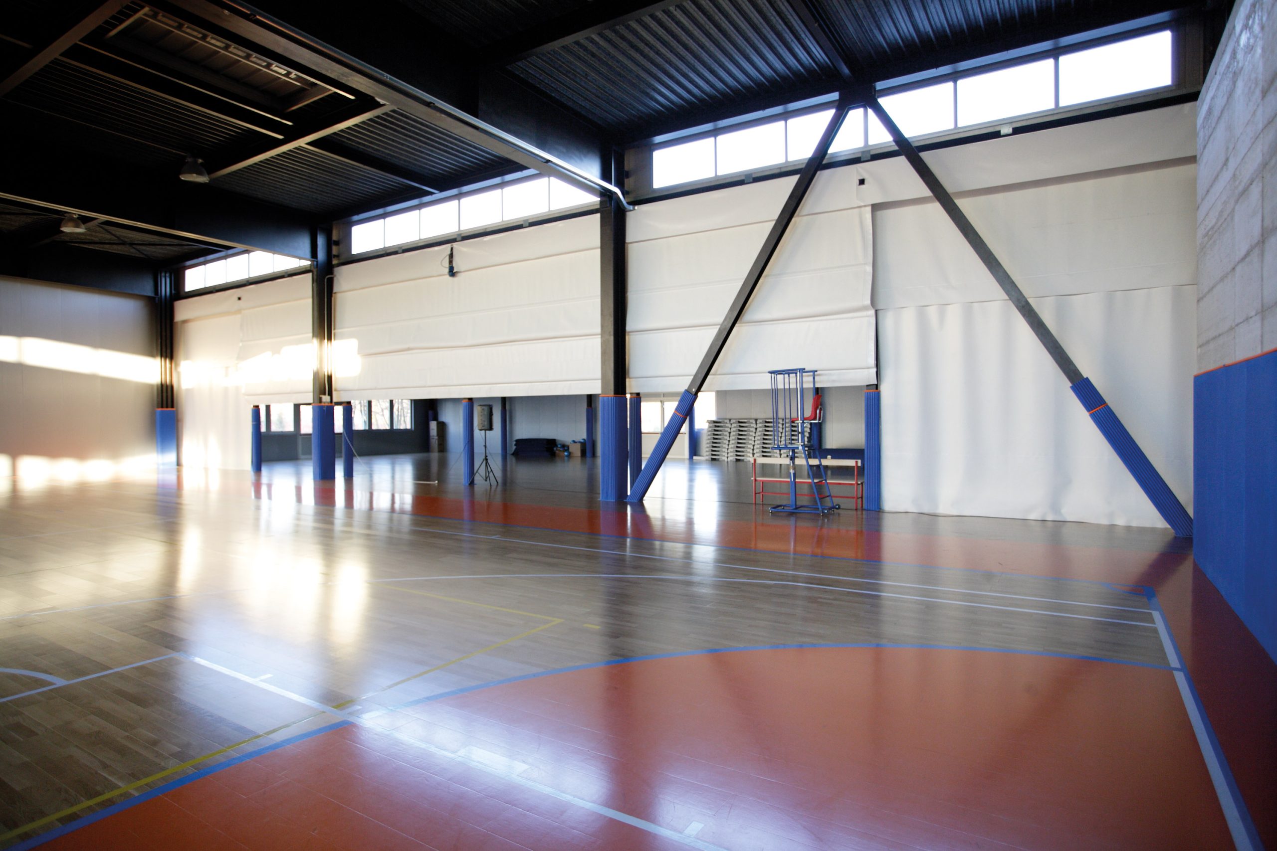 Ampia palestra interna vuota con pavimento in legno, linee di demarcazione blu e arancioni, soffitti alti e ampie finestre che lasciano entrare la luce naturale. Gradini pieghevoli e un canestro da basket sono visibili vicino alla parete di fondo.