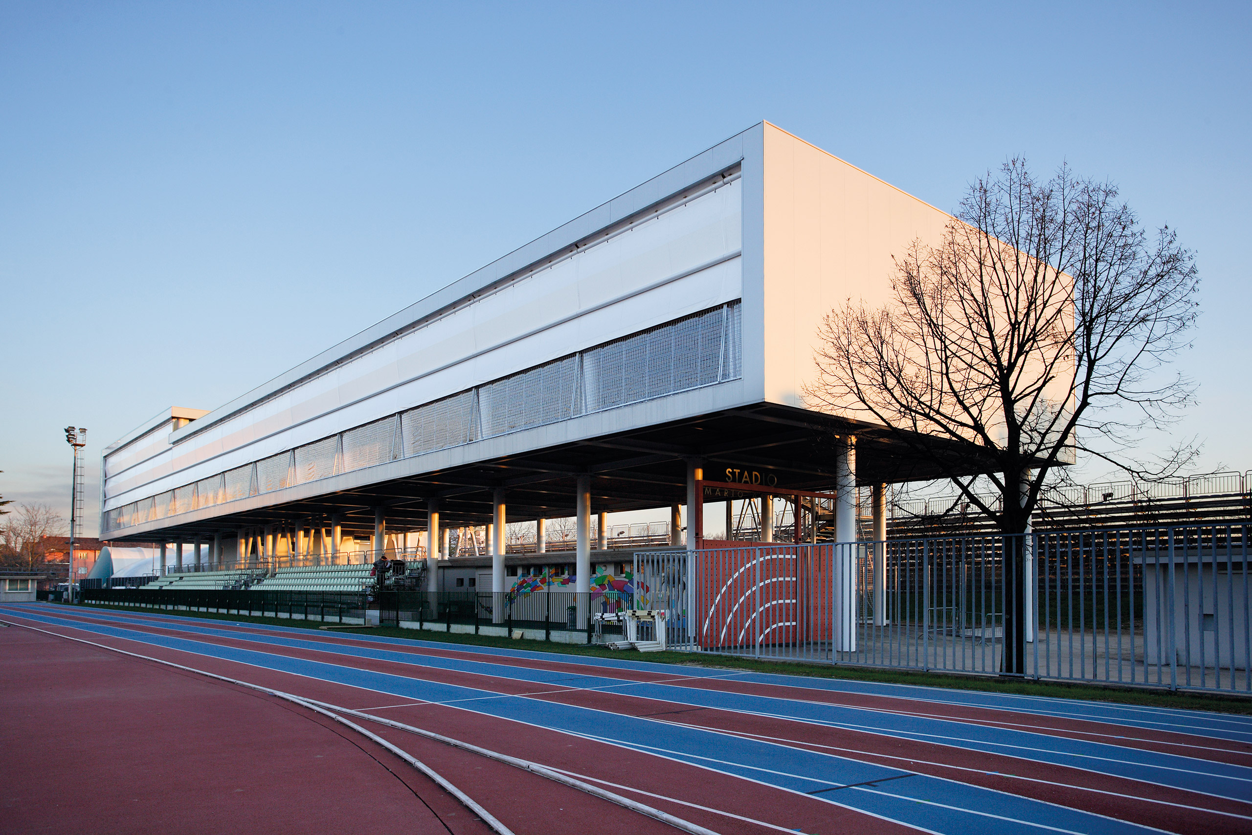 Uno stadio moderno con un elegante edificio rettangolare dotato di posti a sedere aperti nella parte inferiore, accanto a una pista da corsa blu e rossa a più corsie, e con vista sul cielo terso e sugli alberi spogli.