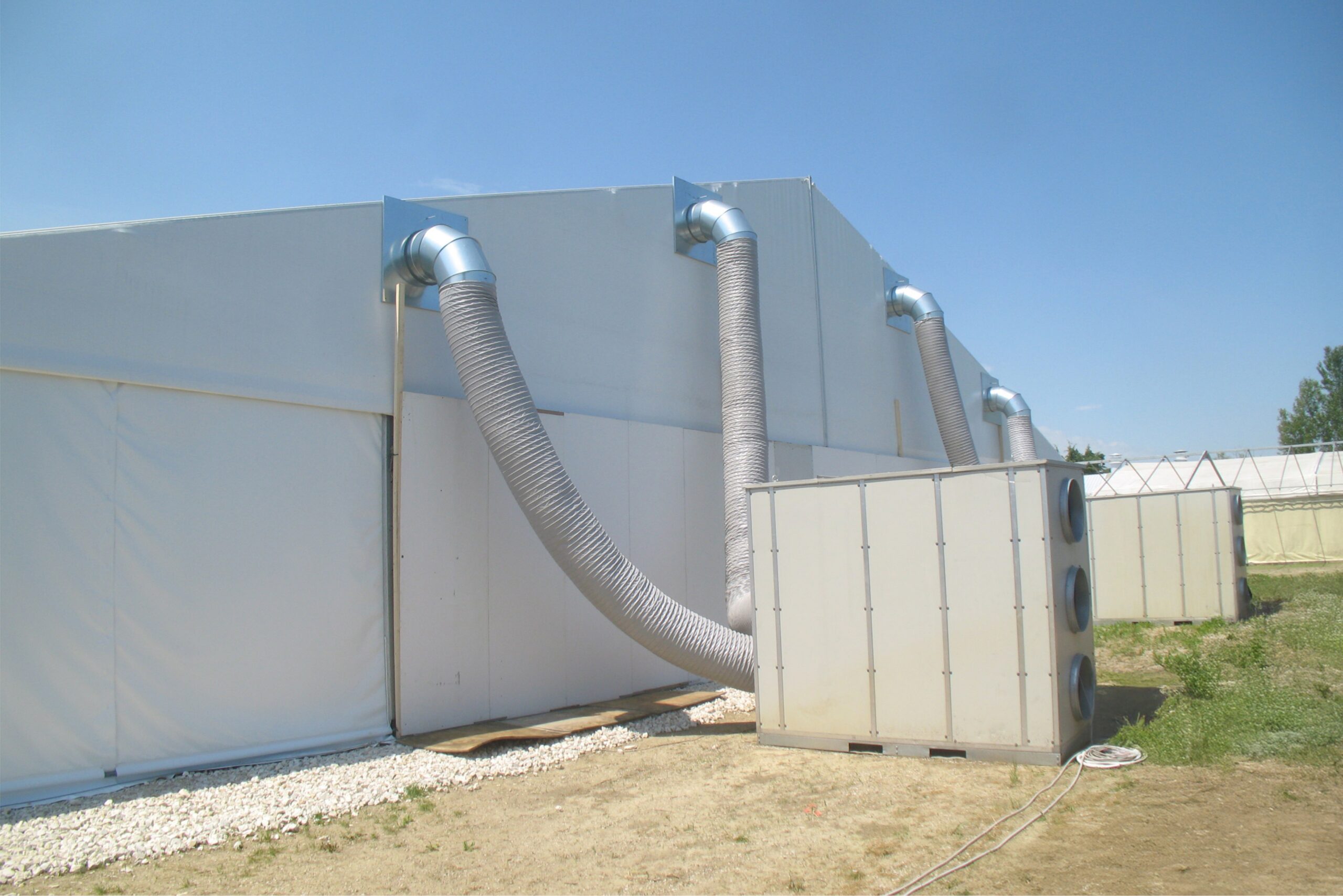 Un grande edificio temporaneo bianco con condotti d'aria flessibili collegati a un'unità HVAC esterna, allestito su un'area erbosa e asciutta sotto un cielo azzurro e limpido.
