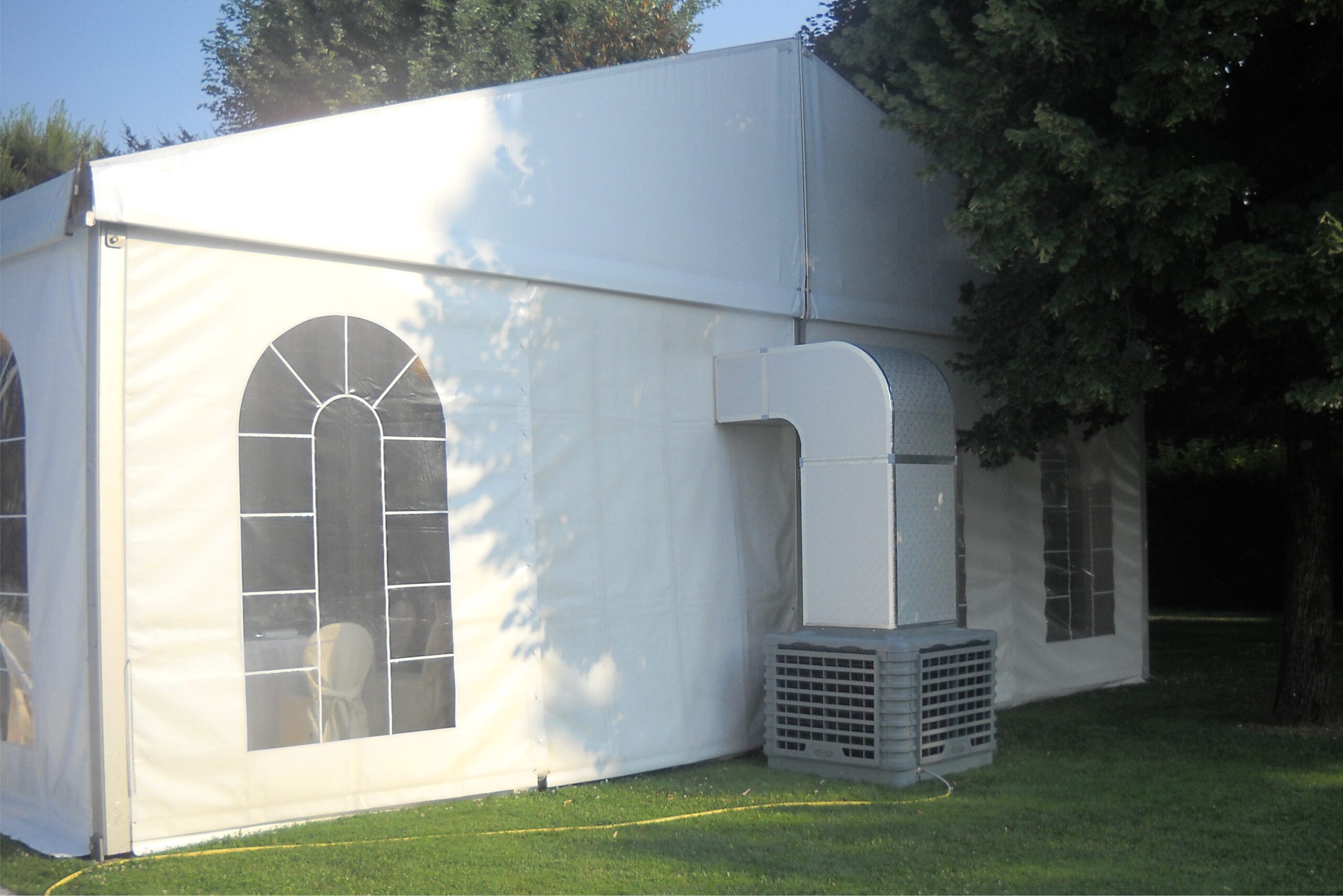 Una grande tenda bianca per eventi all'aperto si trova su un prato erboso, con un'unità di condizionamento e un condotto di ventilazione collegati al lato. Gli alberi forniscono un'ombra parziale accanto alla tenda.