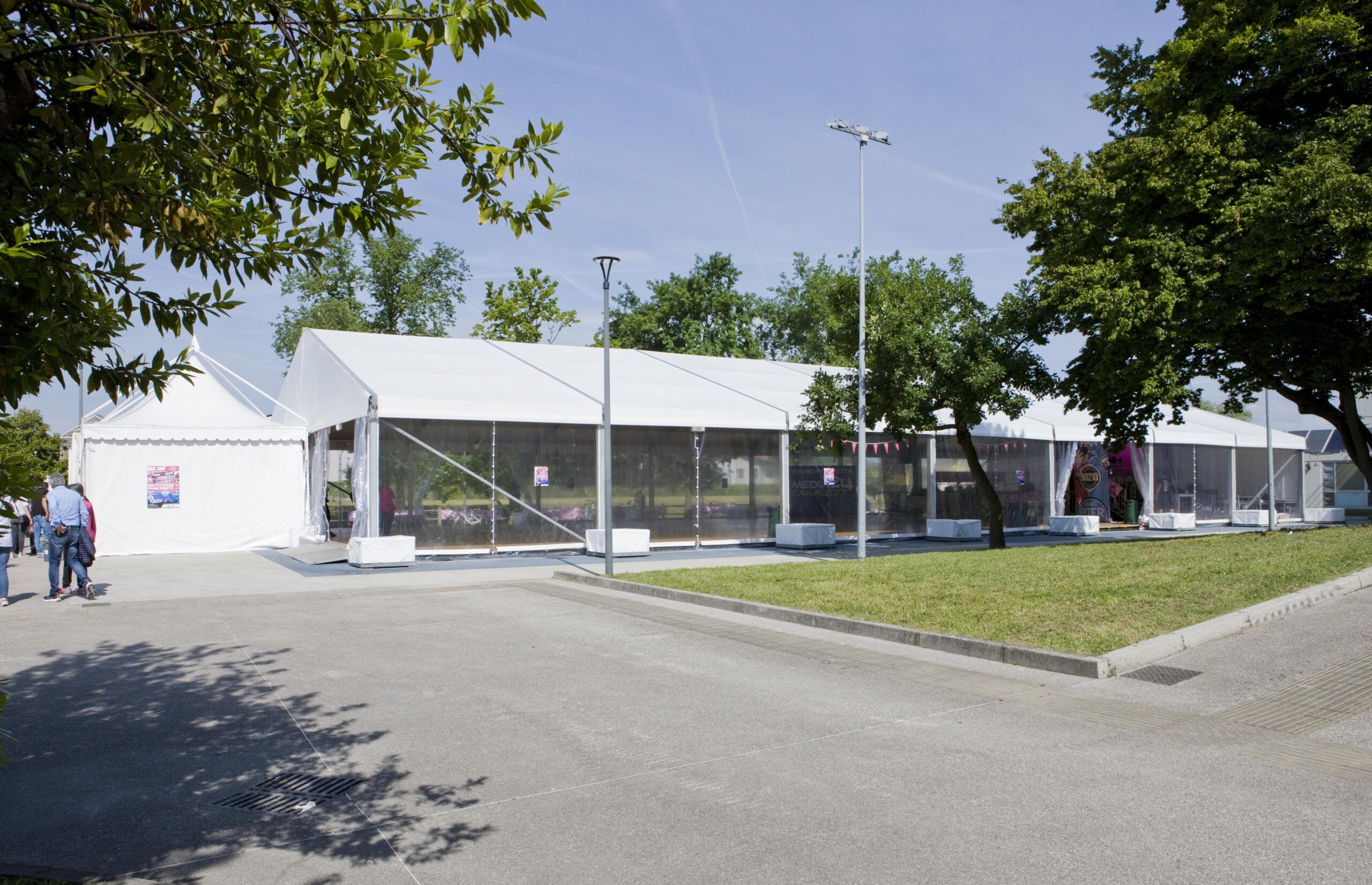 Un grande tendone bianco per eventi con pannelli laterali trasparenti è installato su un'area pavimentata circondata da alberi e prato, sotto un cielo azzurro e terso. Si vedono alcune persone vicino all'ingresso del tendone.