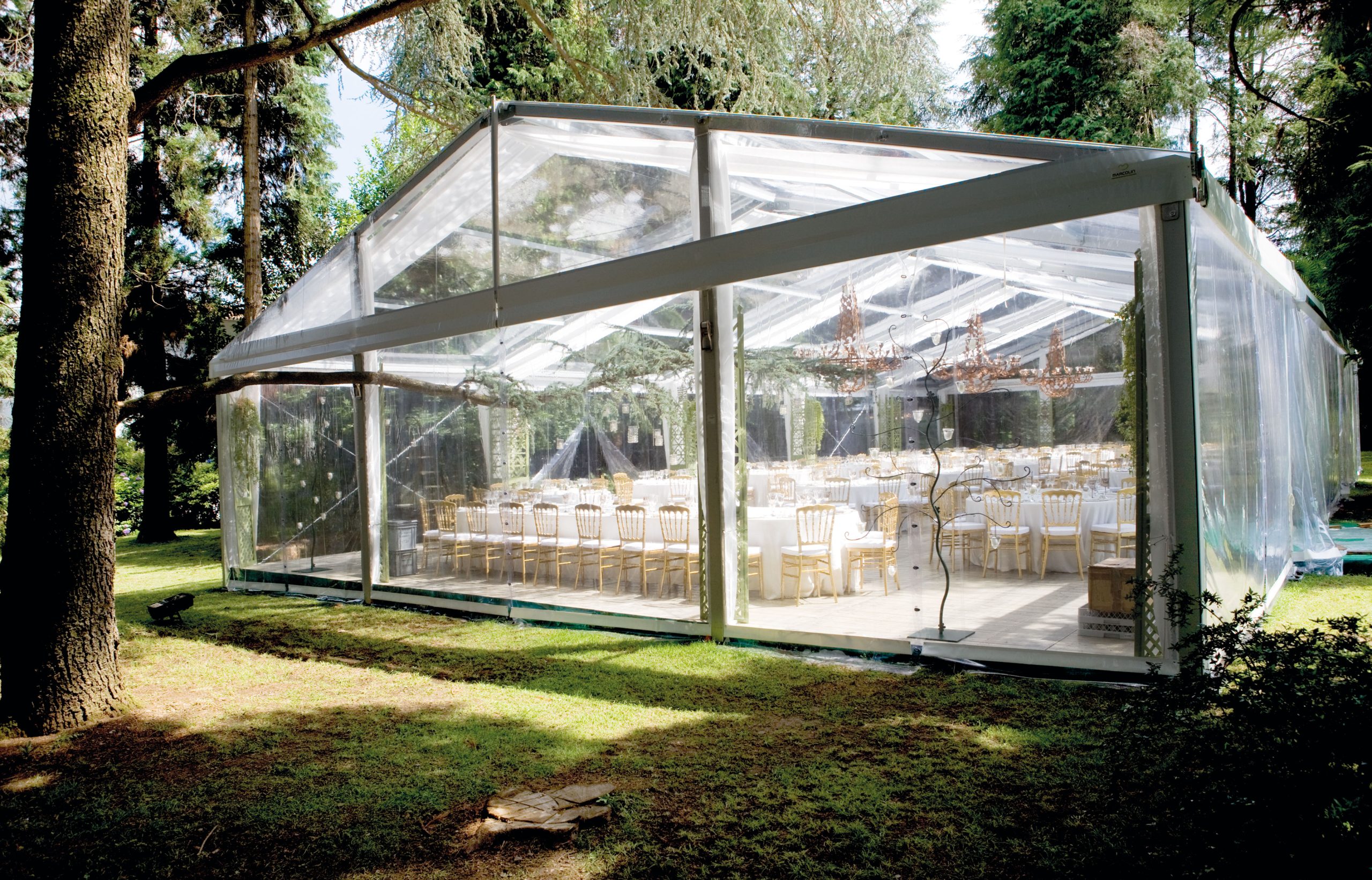 Una grande tenda trasparente allestita all'aperto in una zona boschiva, con file di eleganti tavoli e sedie disposti per un evento o un ricevimento. Lampadari pendono dal soffitto all'interno della tenda.