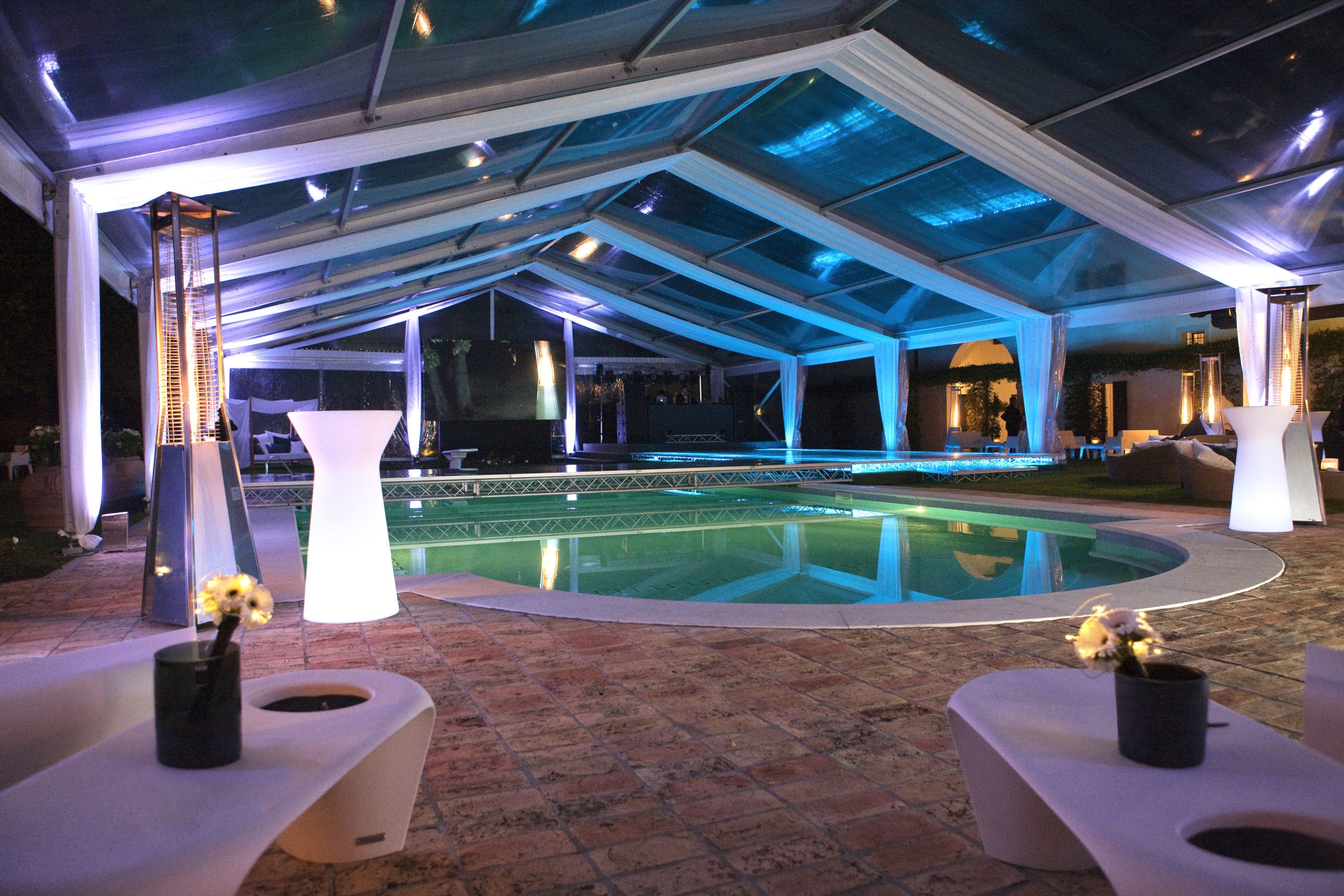 Una lussuosa area piscina interna con soffitto in vetro, elegante illuminazione, moderni mobili bianchi e piccoli fiori in vaso, creano un'atmosfera sofisticata per un evento serale.