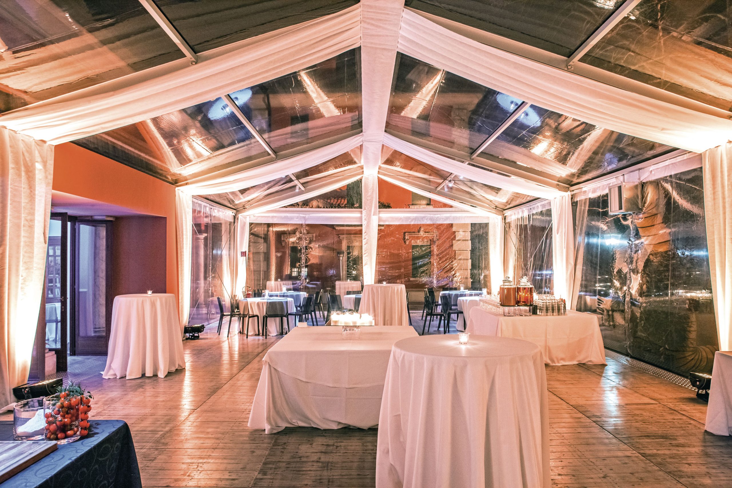 Una tenda trasparente con tessuto drappeggiato e luci a filo copre un'ampia area per eventi allestita con alti tavoli rotondi, tovaglie bianche e candele, creando un'atmosfera elegante e soffusa per un incontro.