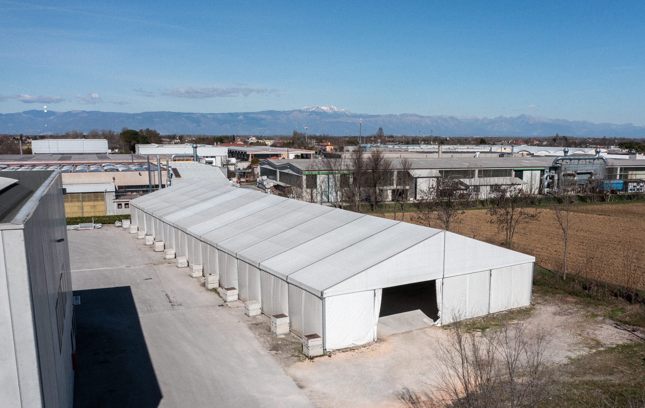 In una giornata limpida, tra gli edifici industriali viene allestita una grande tenda bianca, con i campi nelle vicinanze e le montagne innevate visibili sullo sfondo in lontananza.