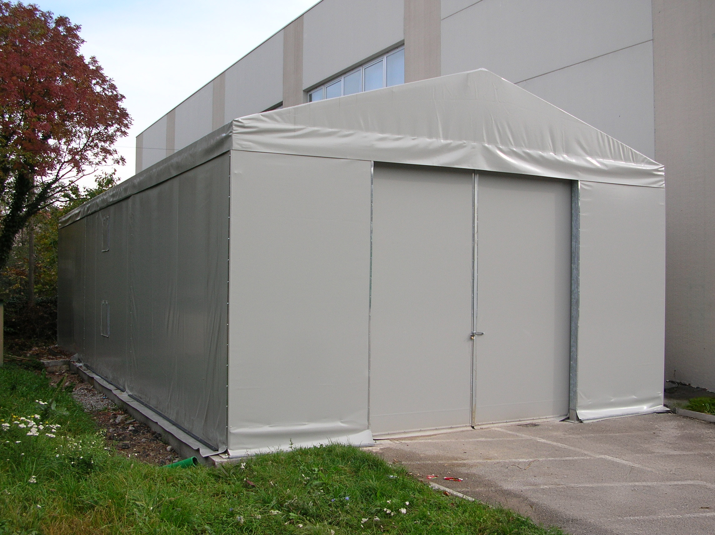 Una grande tenda temporanea grigio chiaro con porte metalliche è posta su una piattaforma di cemento accanto a un moderno edificio industriale, con erba e un albero nelle vicinanze.