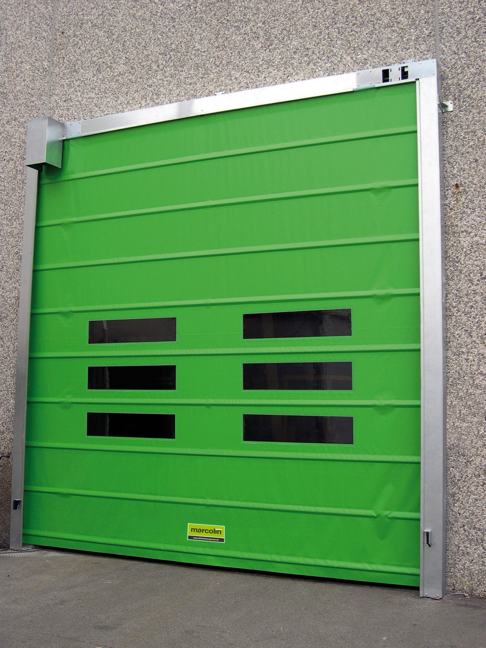 Una porta industriale avvolgibile verde brillante con tre finestre rettangolari orizzontali, inserita in un muro grigio con texture in cemento. Il telaio della porta è metallico e una targhetta con il logo giallo è visibile in basso al centro.