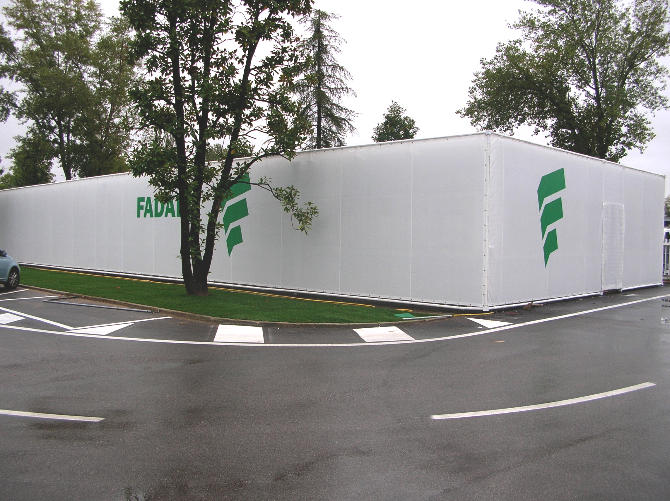 Un grande edificio temporaneo bianco con loghi verdi sui lati si erge su un marciapiede bagnato, circondato da alberi e prato. Un'auto parcheggiata è parzialmente visibile sulla sinistra.