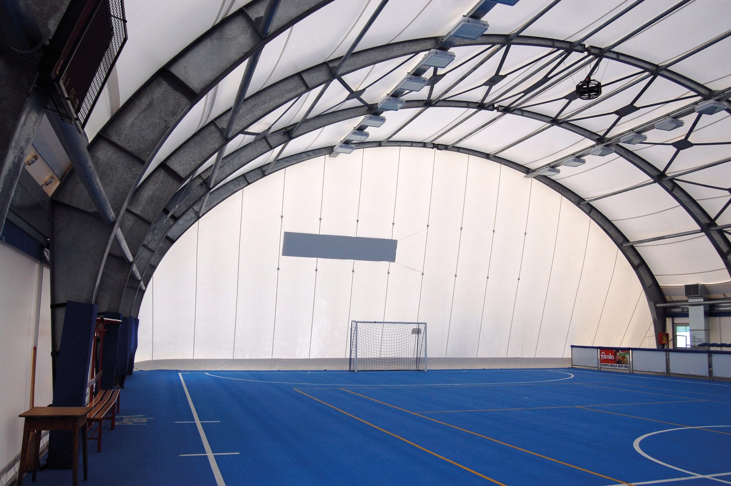 Un campo sportivo coperto con pavimento blu, linee bianche e una porta da calcio a un'estremità. Il tetto ad arco è realizzato in materiale bianco traslucido, che lascia entrare la luce naturale. Lo spazio è vuoto, con panchine e un tavolo sulla sinistra.