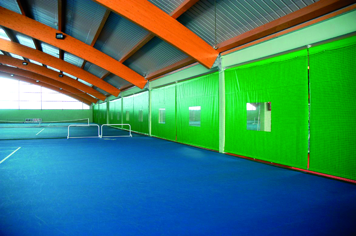 Campo da tennis coperto con pavimento blu, pareti verdi e divisori in rete, soffitto alto con travi in legno a vista e luce naturale che entra dalle finestre.