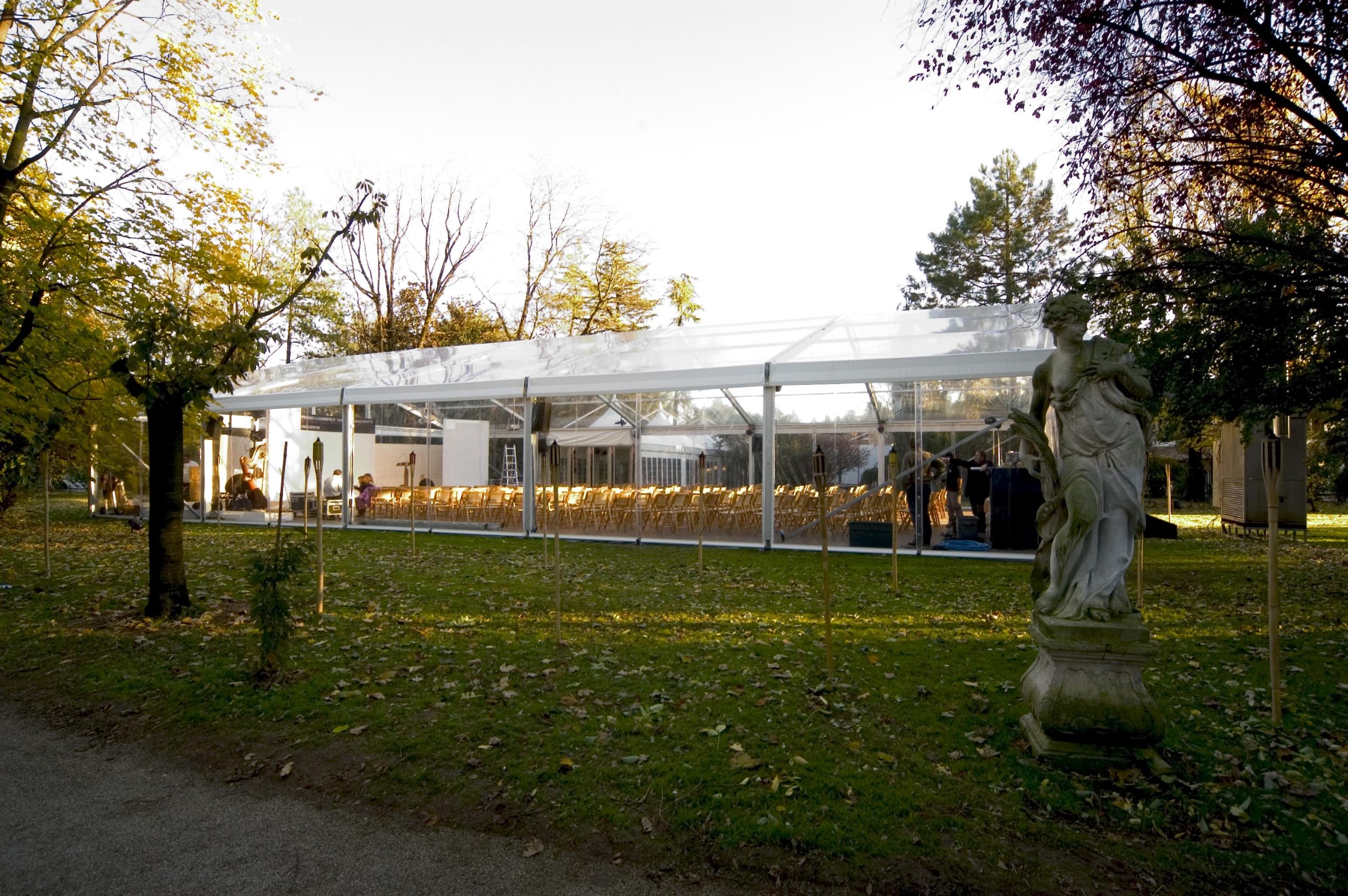 Su un prato erboso circondato da alberi si erge una tenda trasparente con file di sedie vuote al suo interno, con in primo piano una statua classica in pietra.