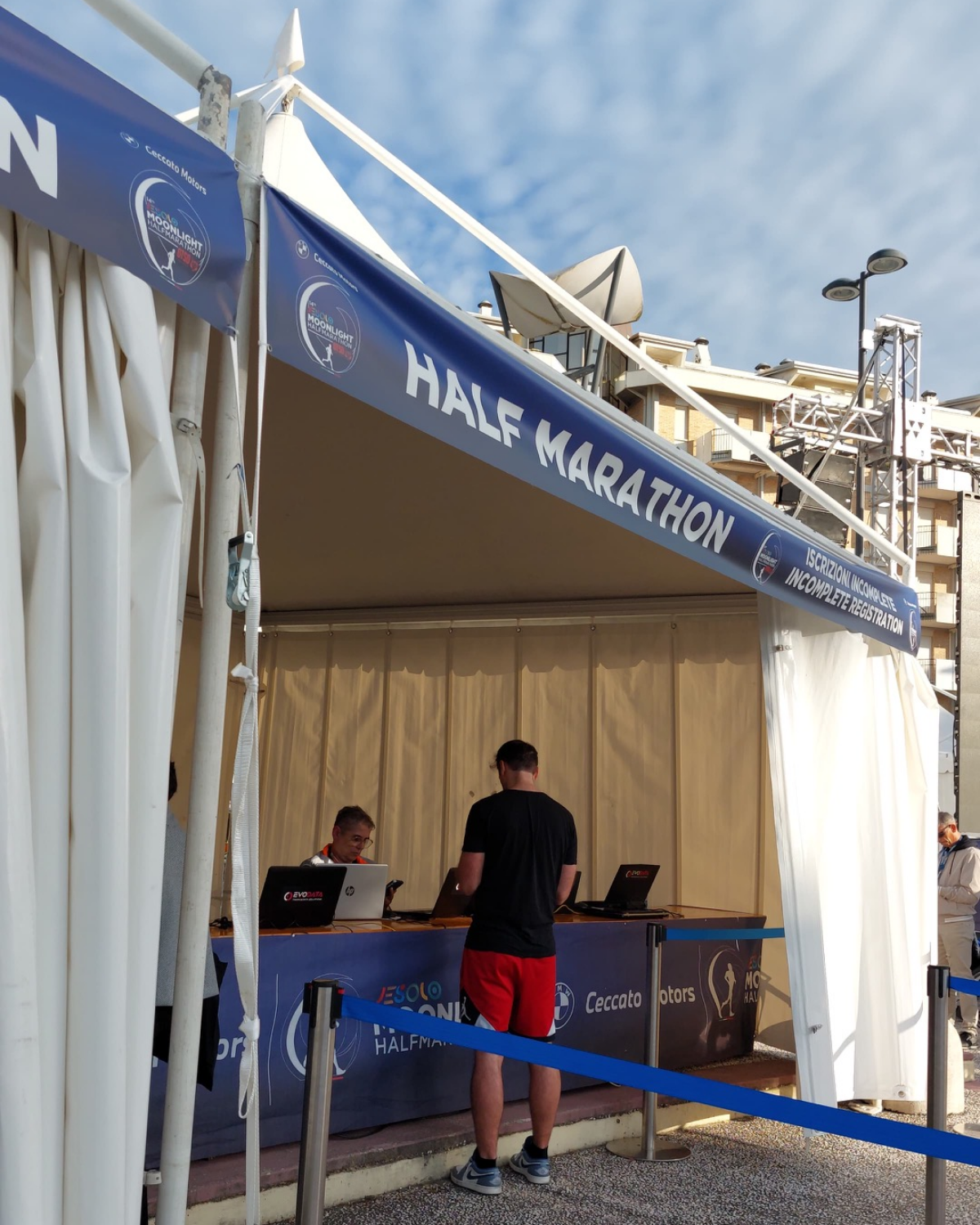 Un uomo è in piedi davanti allo stand di registrazione, sotto una tettoia con la scritta "MEZZA MARATONA", e parla con il personale seduto a tavoli con computer portatili; sullo sfondo sono visibili edifici e un cielo parzialmente nuvoloso.
