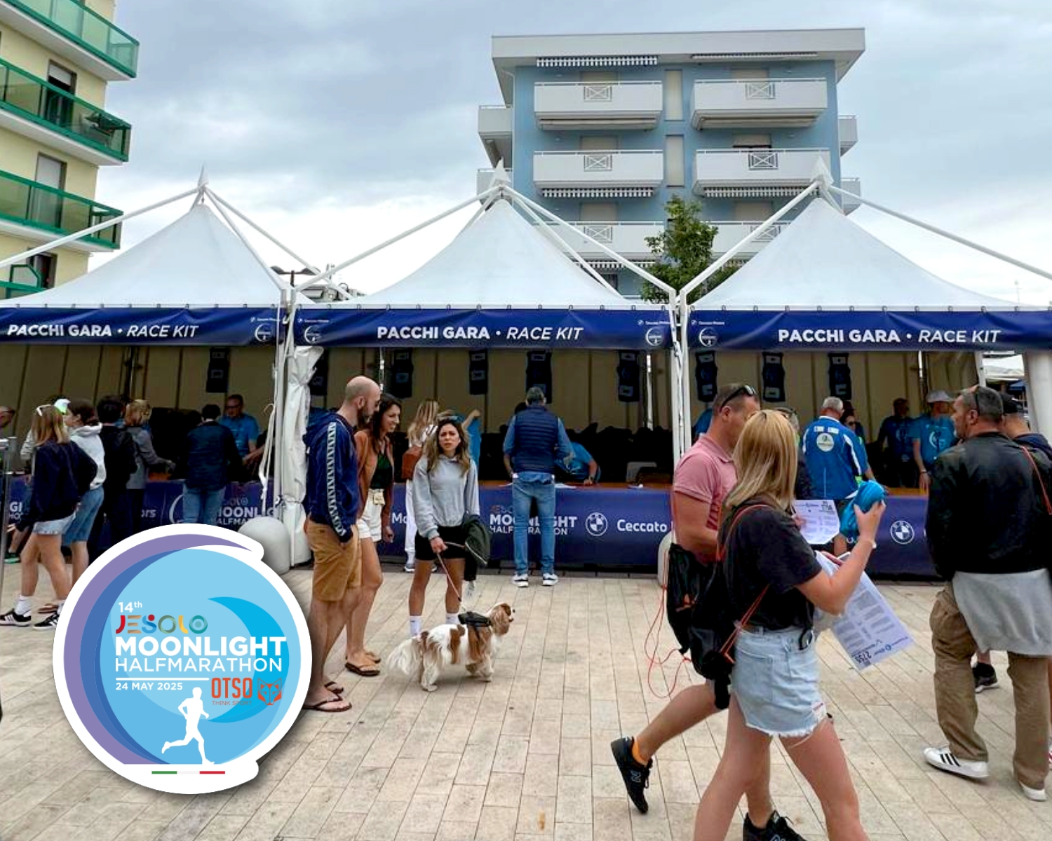 La gente si raduna davanti alle tende bianche con la scritta "PACCHI GARA - RACE KIT" alla Jesolo Moonlight Half Marathon. Alcuni portano a spasso i cani, altri controllano i documenti. Sono visibili gli edifici vicini e il logo dell'evento.