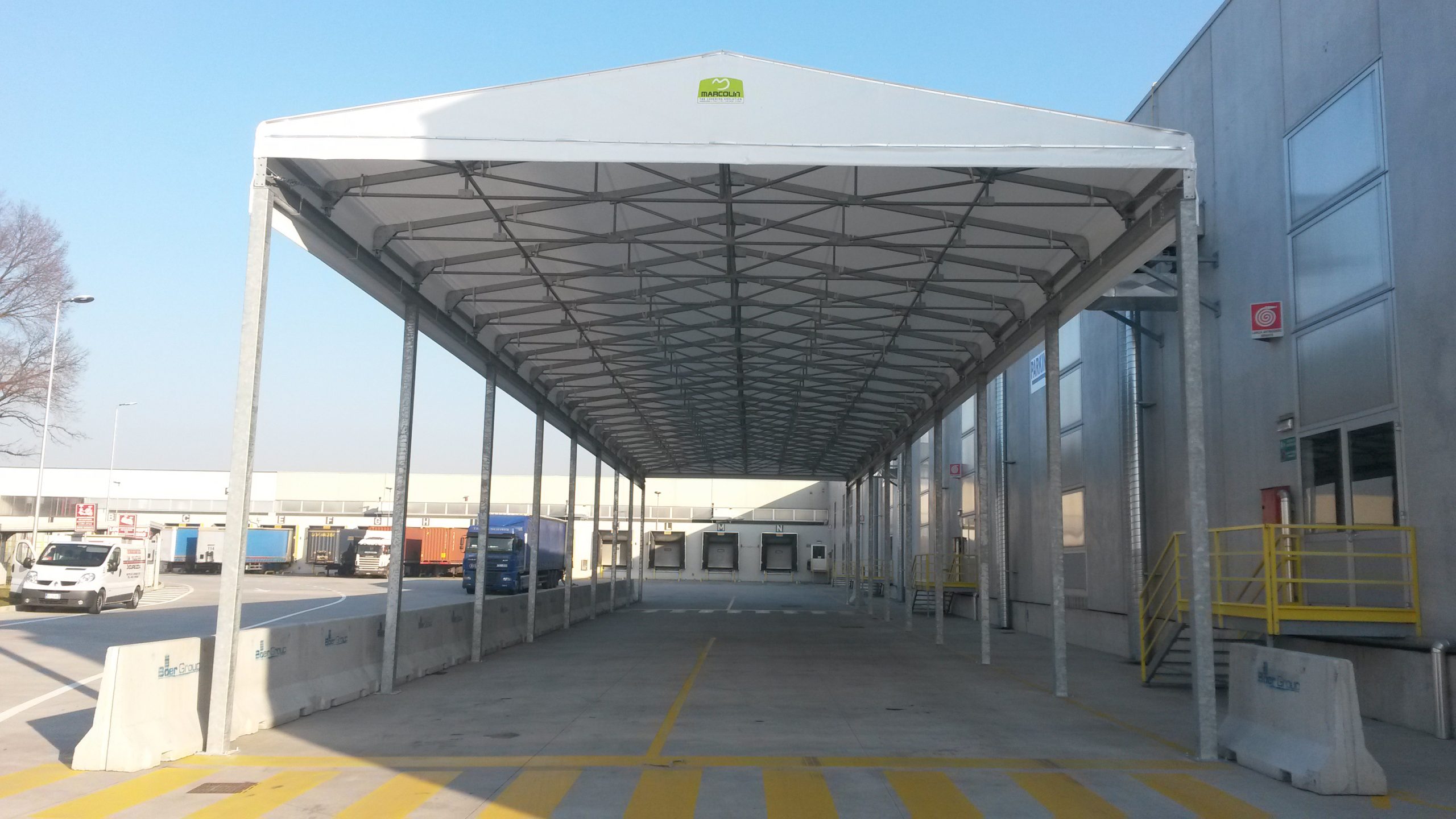 Una grande struttura bianca con tettoia aperta lateralmente si erge accanto a un edificio industriale con scale gialle, offrendo uno spazio coperto sopra un'area di carico in cemento. Sullo sfondo sono visibili diversi camion e veicoli.