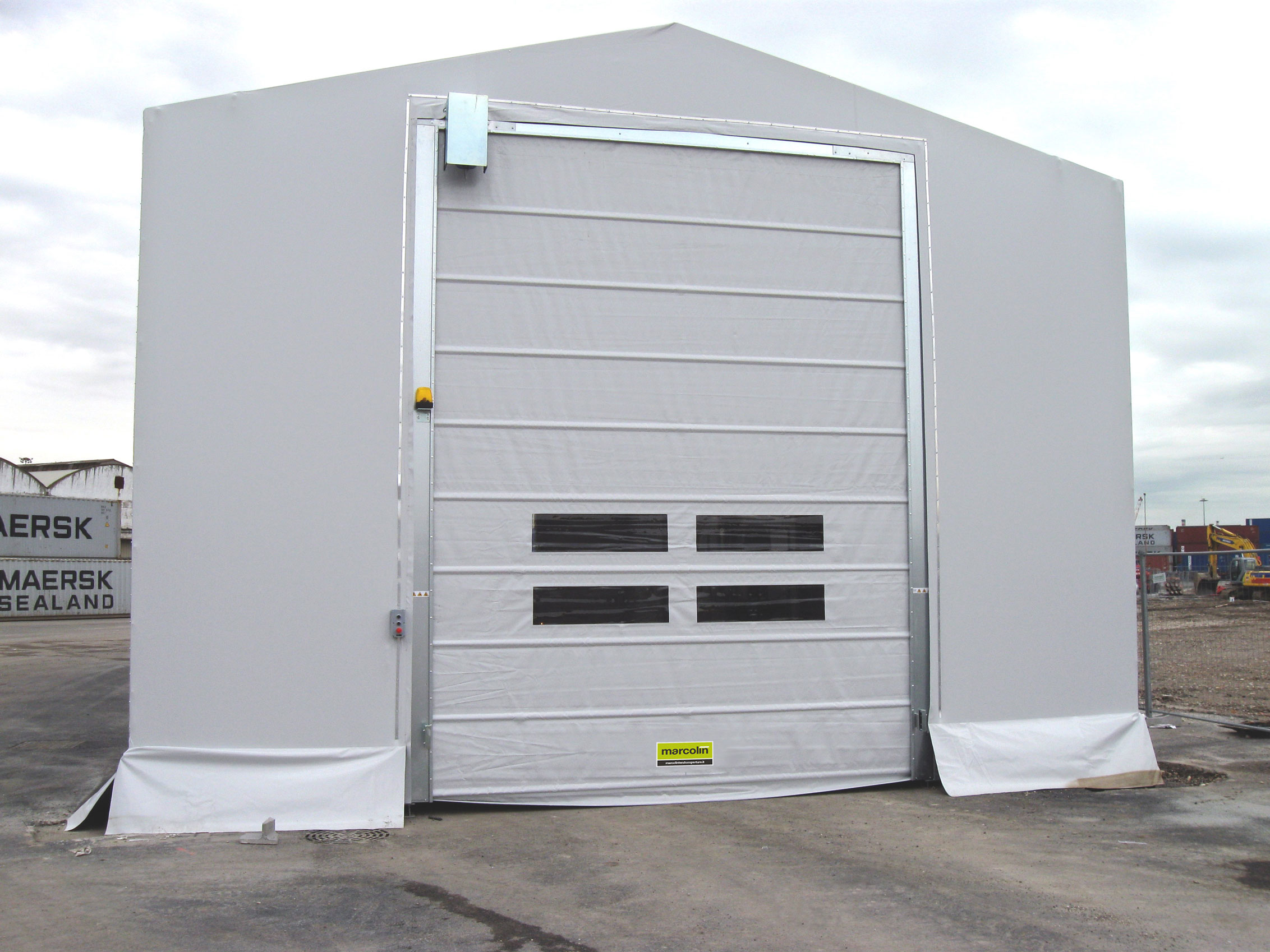 Una grande tenda industriale bianca con una porta avvolgibile con telaio metallico e due finestre orizzontali, posizionata su una superficie pavimentata in un cantiere o in un deposito.