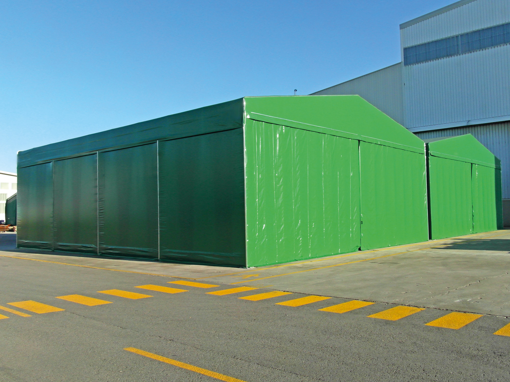Grande struttura temporanea verde o tenda con lati chiusi, situata su una superficie pavimentata all'esterno di un edificio industriale, sotto un cielo azzurro e limpido. In primo piano sono visibili le linee gialle di parcheggio o di traffico.