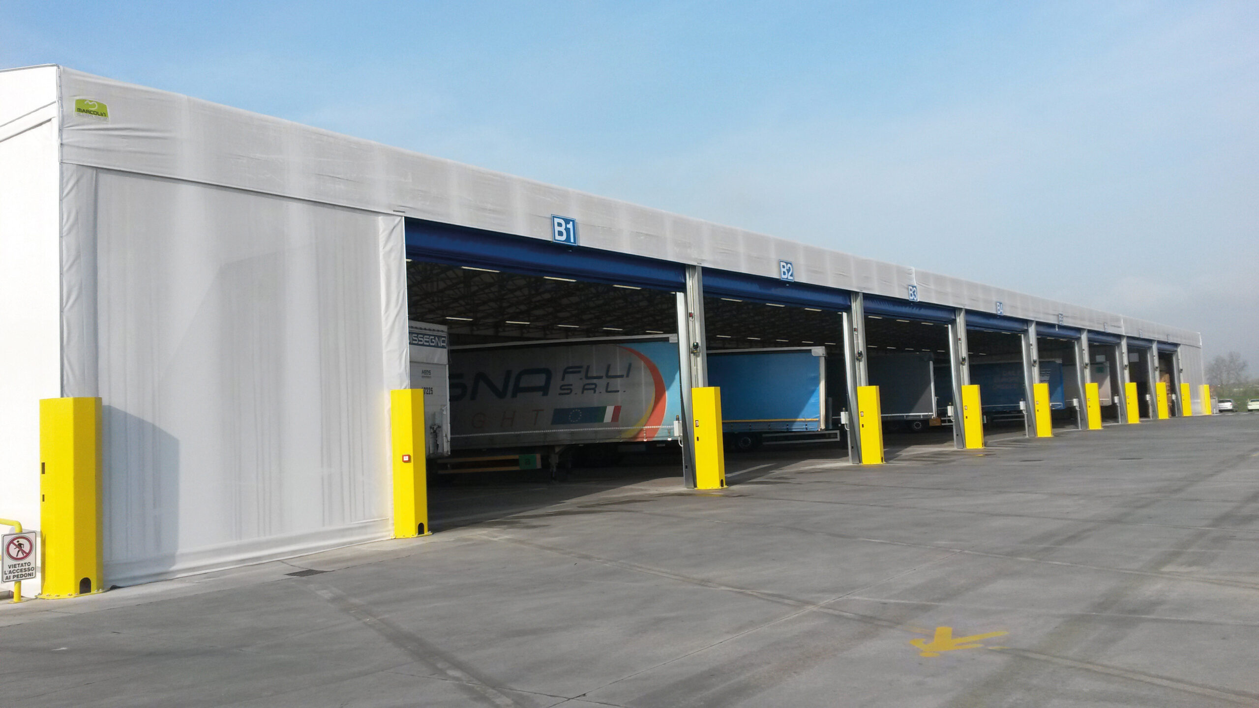 Una grande banchina di carico industriale con diverse baie aperte, bitte di sicurezza gialle e un rimorchio di camion parcheggiato all'interno di una baia. L'area è pulita e il cielo è limpido.