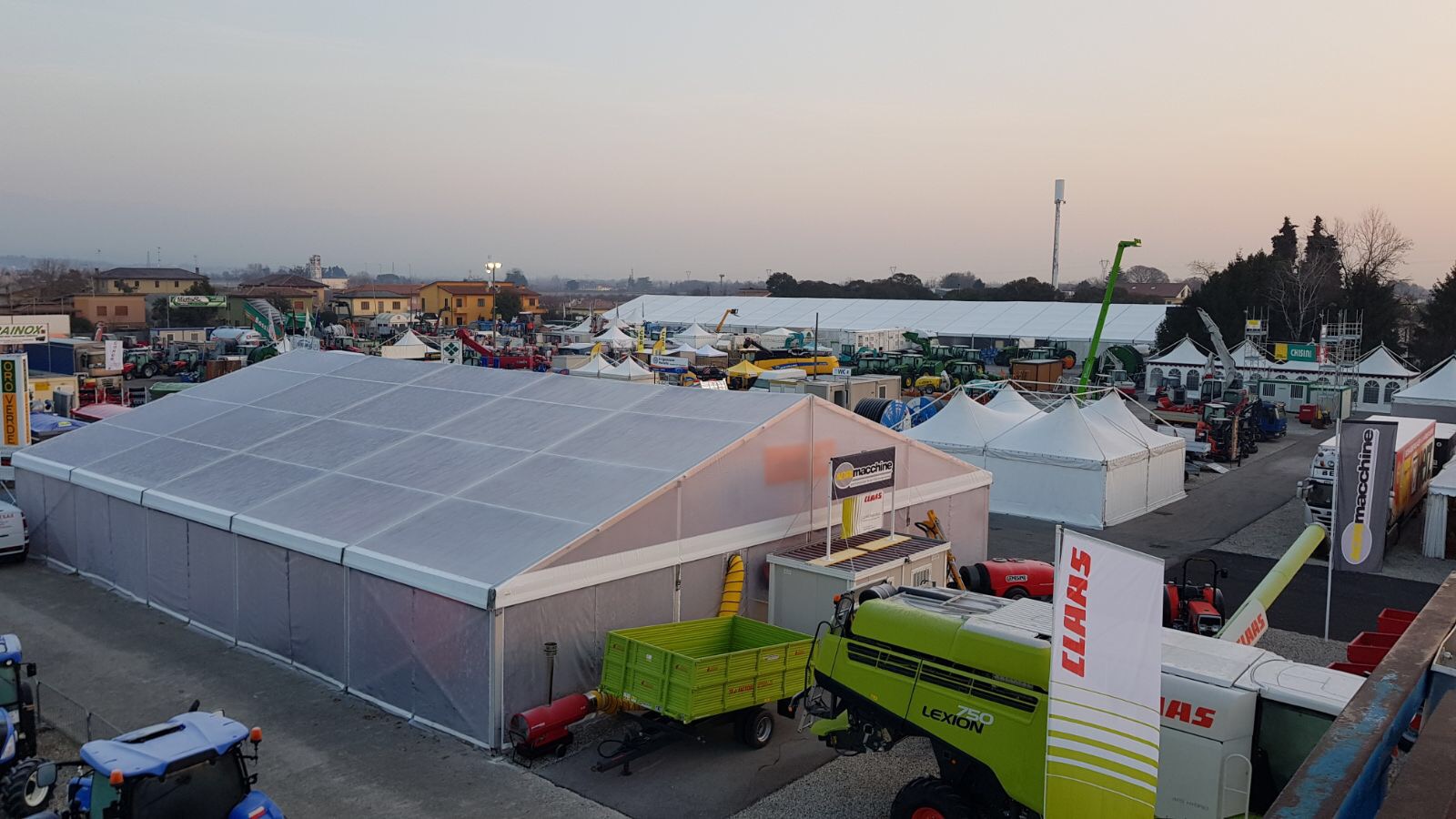 Grande fiera agricola all'aperto con tende, macchinari e attrezzature agricole in esposizione. Diversi trattori, rimorchi e stand con i marchi sono visibili sotto un cielo terso al tramonto.