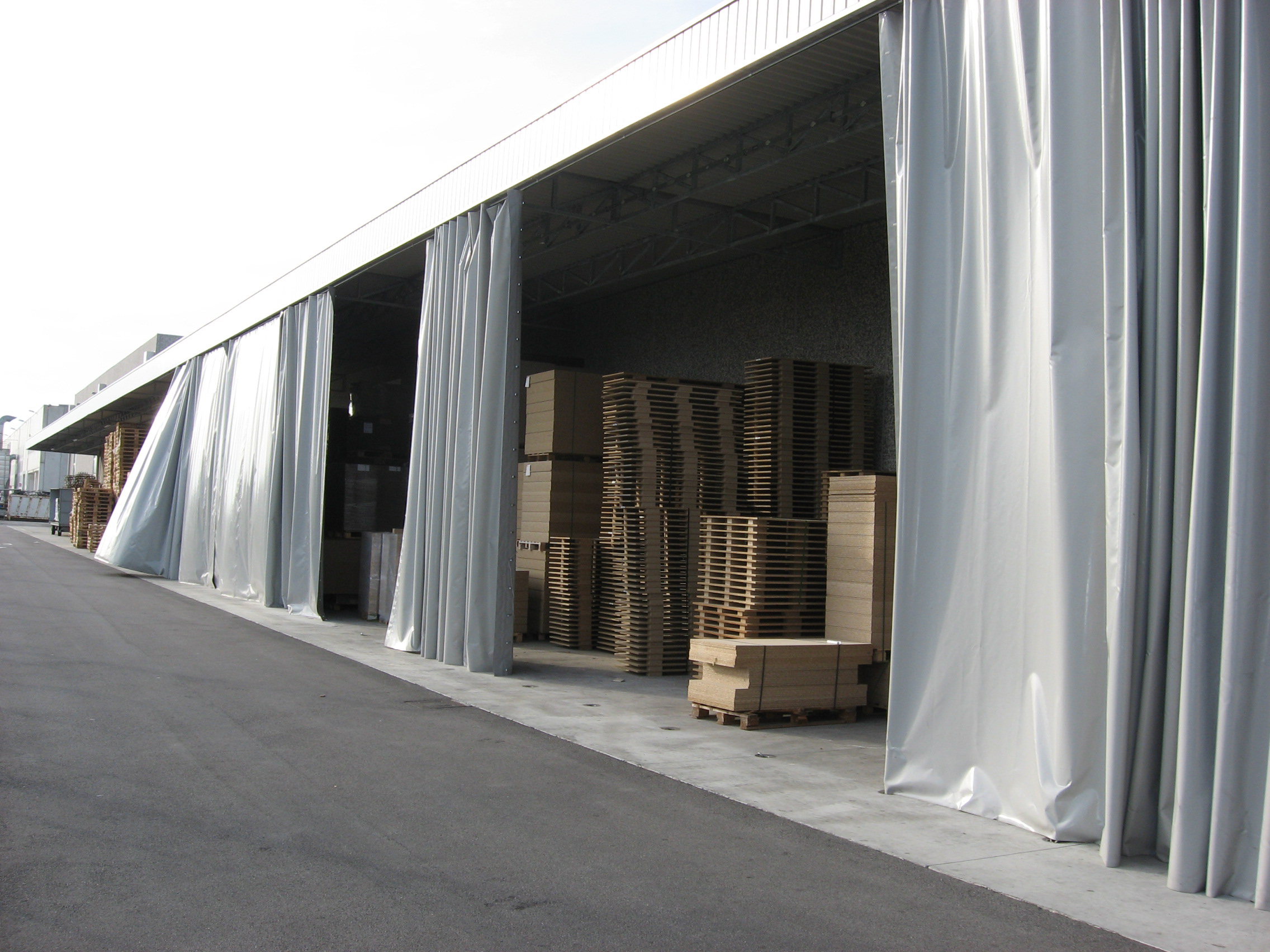 Pile di pallet di legno sono immagazzinate all'interno di un magazzino industriale con grandi porte argentate, simili a tende, parzialmente aperte all'esterno.