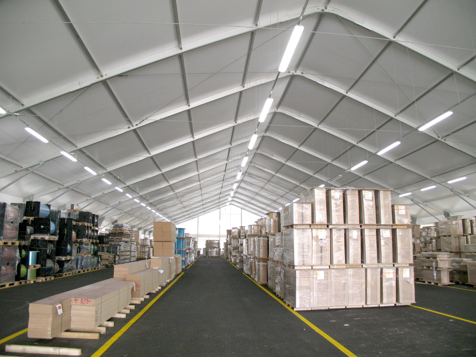 Interno di un grande magazzino con soffitti alti, pieno di pallet e scatole ordinatamente accatastati lungo entrambi i lati di ampie corsie centrali, sotto una forte illuminazione dall'alto.