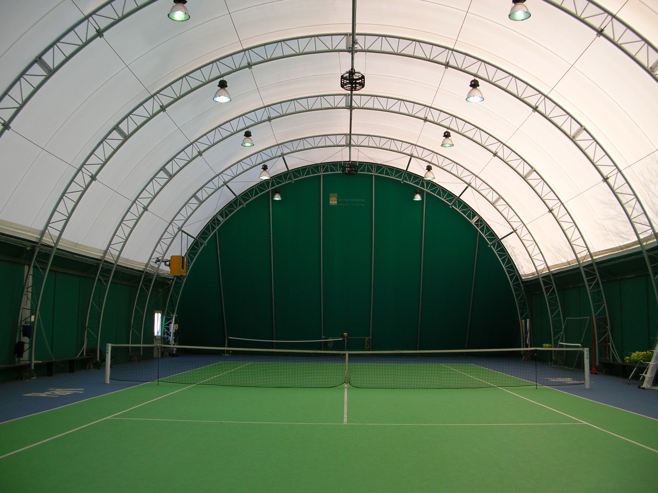 Un campo da tennis coperto con superficie verde e blu, circondato da alte pareti verdi e coperto da un'ampia copertura curva in tela bianca con supporti metallici e luci a soffitto. La rete è posizionata al centro.