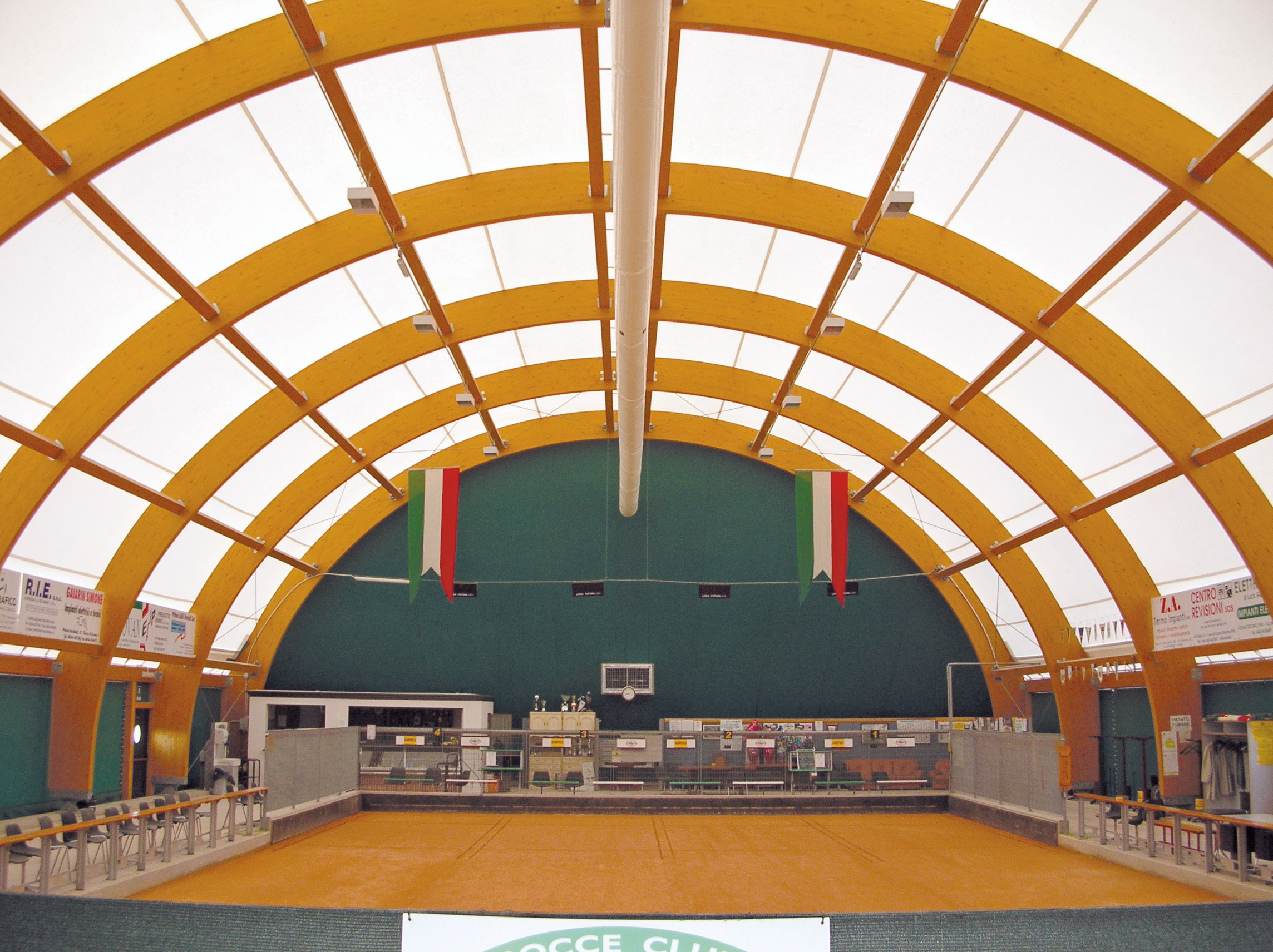 Interno di un impianto sportivo coperto con soffitto con travi di legno curve, pavimento arancione, due bandiere ungheresi appese, panchine lungo i lati e trofei esposti all'estremità più lontana.