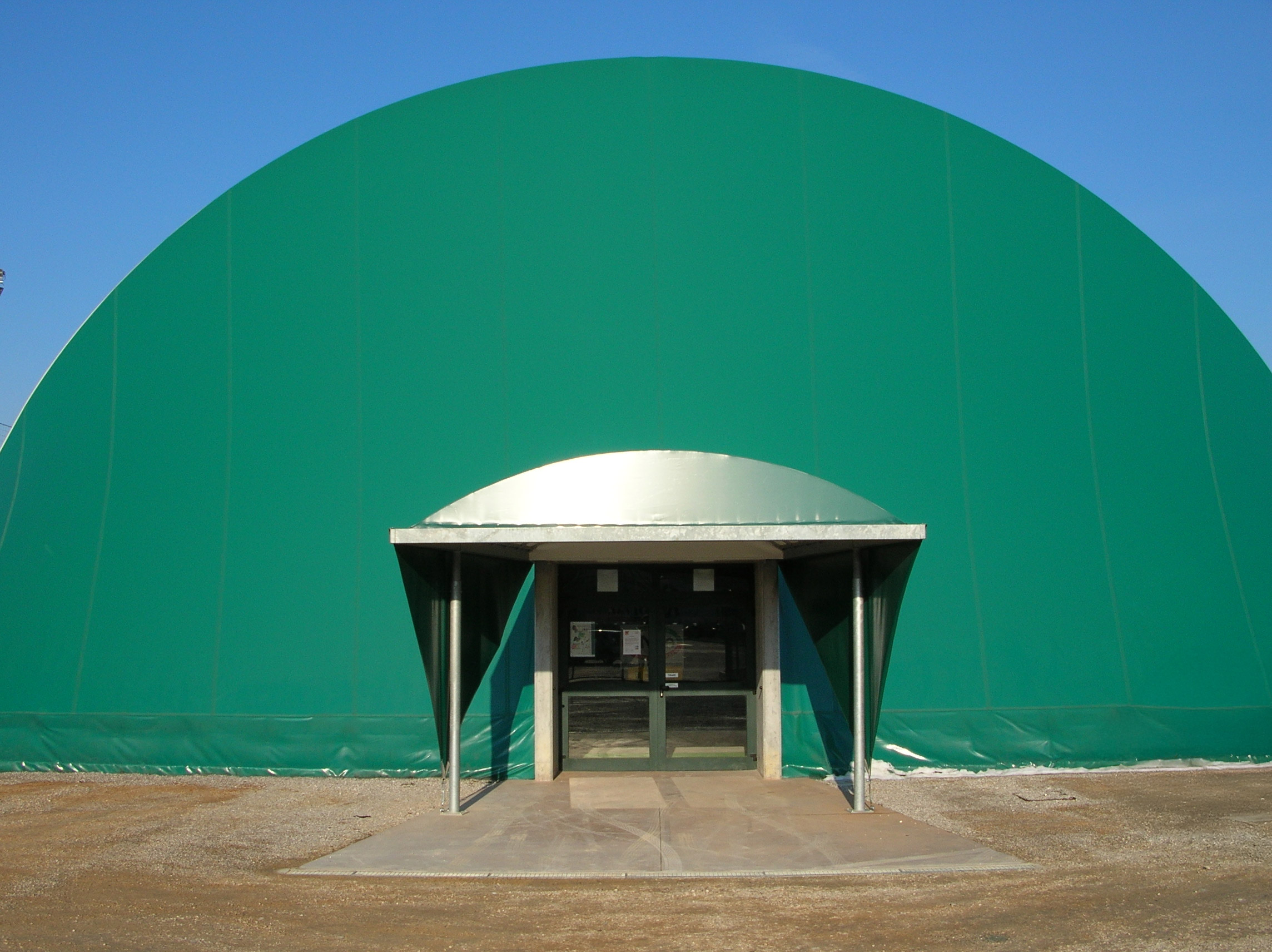 Un grande edificio verde a forma di cupola con un ingresso centrale caratterizzato da porte in vetro e una piccola pensilina metallica, che si staglia contro un cielo azzurro terso. Il terreno antistante è pavimentato e delimitato da ghiaia.