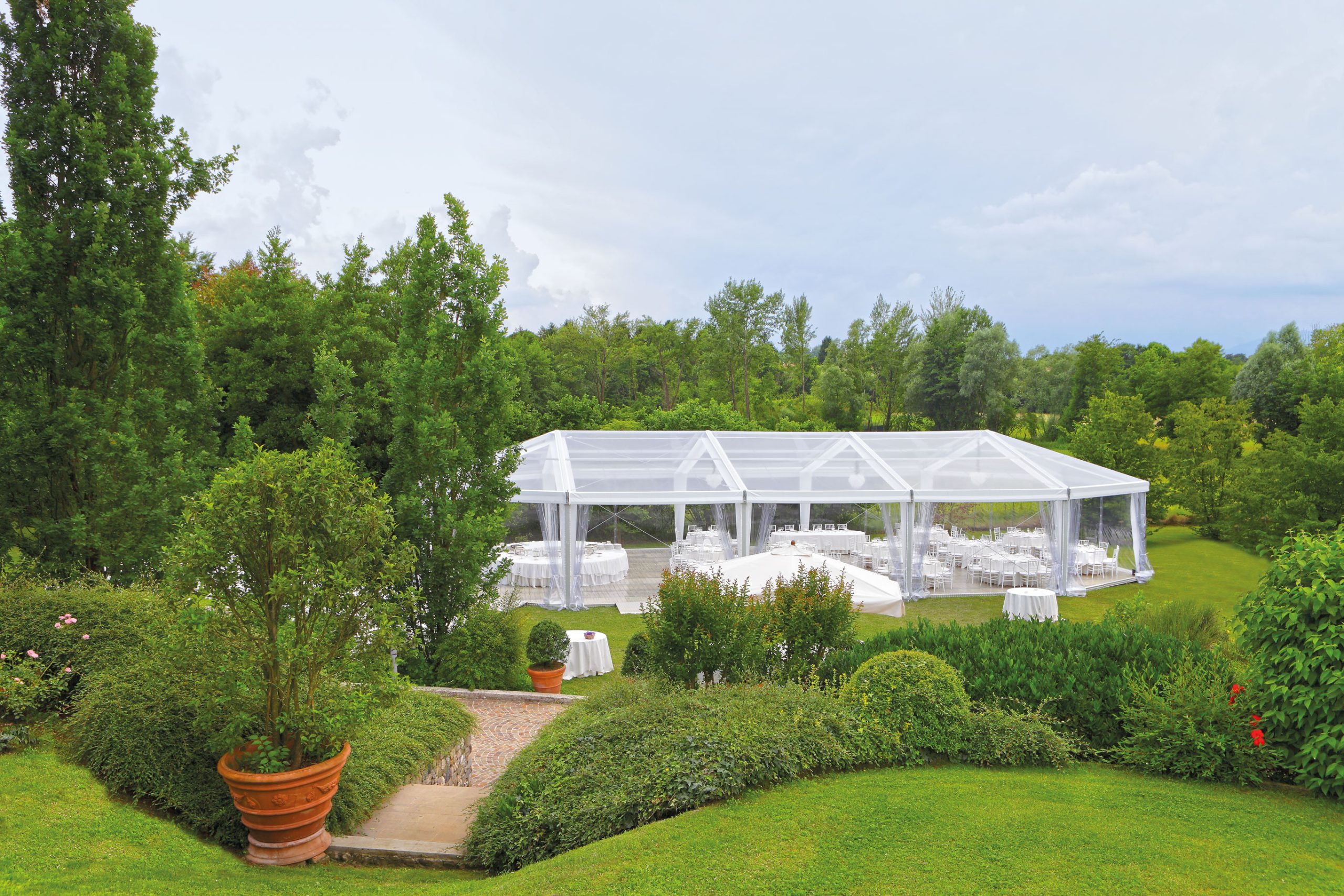 Una tenda trasparente allestita per un evento all'aperto è adagiata su un prato ben curato, circondata da rigogliosi alberi verdi, cespugli e piante da giardino, sotto un cielo parzialmente nuvoloso.