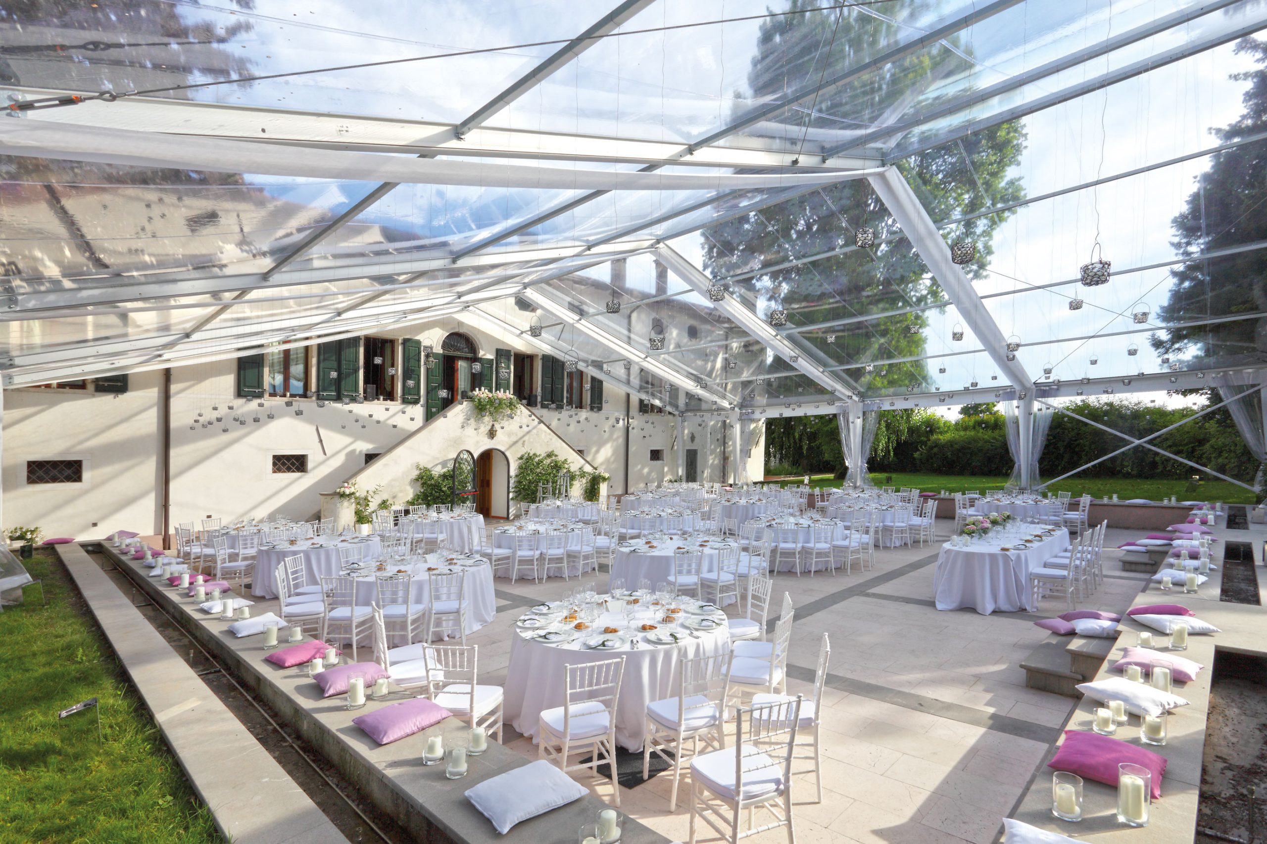 Un ampio ricevimento di nozze all'aperto allestito sotto una tenda trasparente con tavoli rotondi e rettangolari coperti da tovaglie bianche, sedie bianche e cuscini color lavanda, circondato dal verde e con un grande edificio sullo sfondo.