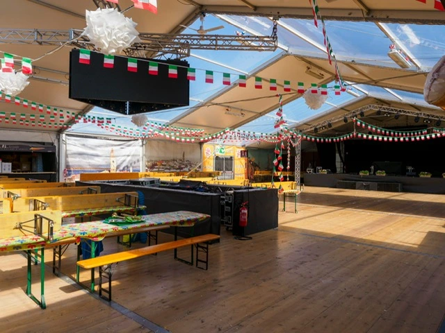 Una grande tenda decorata con festoni con la bandiera italiana e lanterne di carta, con lunghi tavoli e panche in legno, allestita per un evento con pavimento in legno e tetto trasparente che lascia entrare la luce del sole.