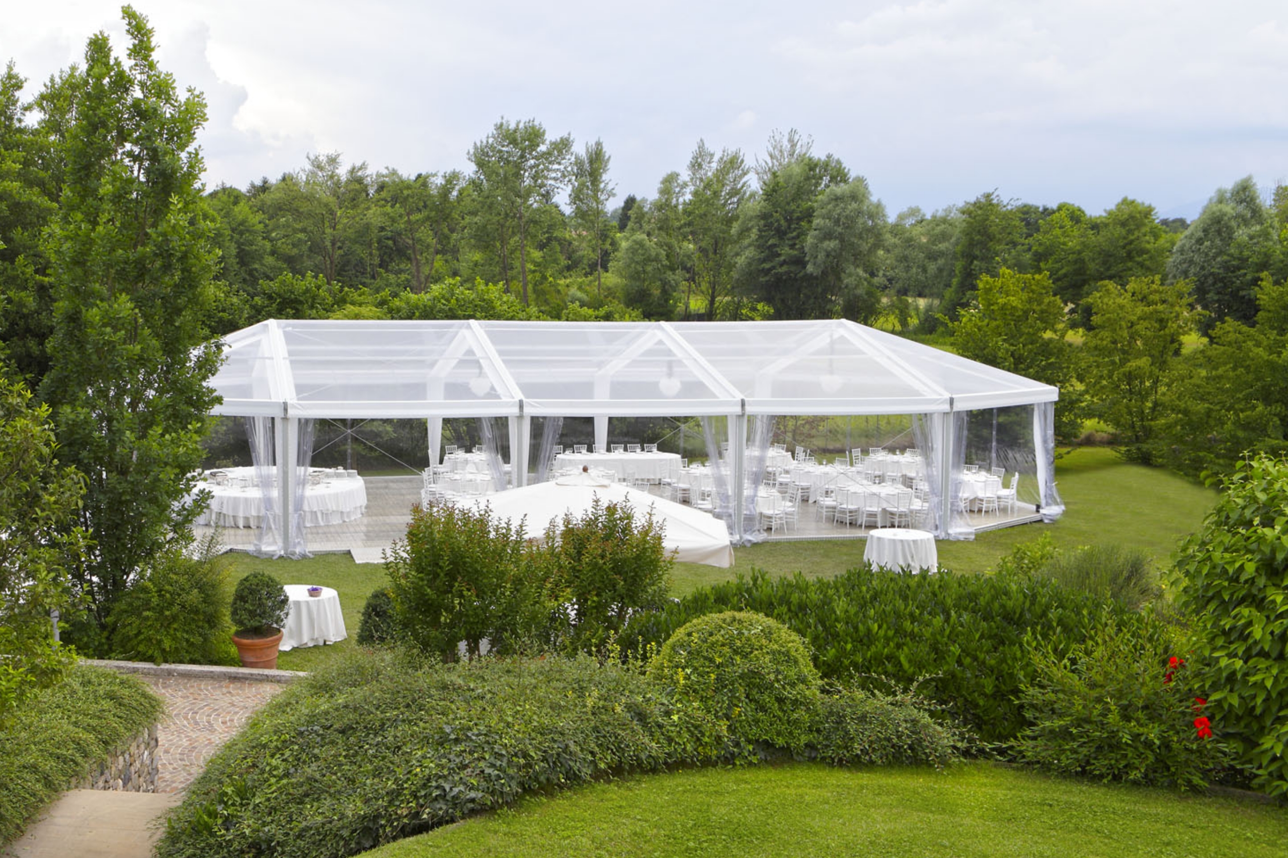 Per l'evento viene allestita una tenda trasparente posizionata su un prato erboso, circondata da rigogliosi alberi e cespugli verdi, con tavoli rotondi e sedie bianche disposti ordinatamente al suo interno.
