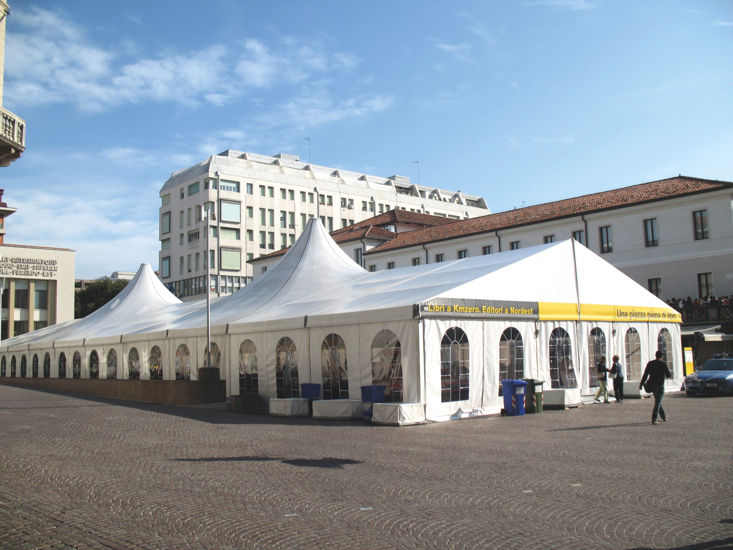 Un grande tendone bianco con la cima a punta è allestito su una piazza acciottolata, circondato da edifici moderni e tradizionali sotto un cielo azzurro e terso. La gente passeggia nelle vicinanze e gli striscioni decorano il bordo del tetto del tendone.