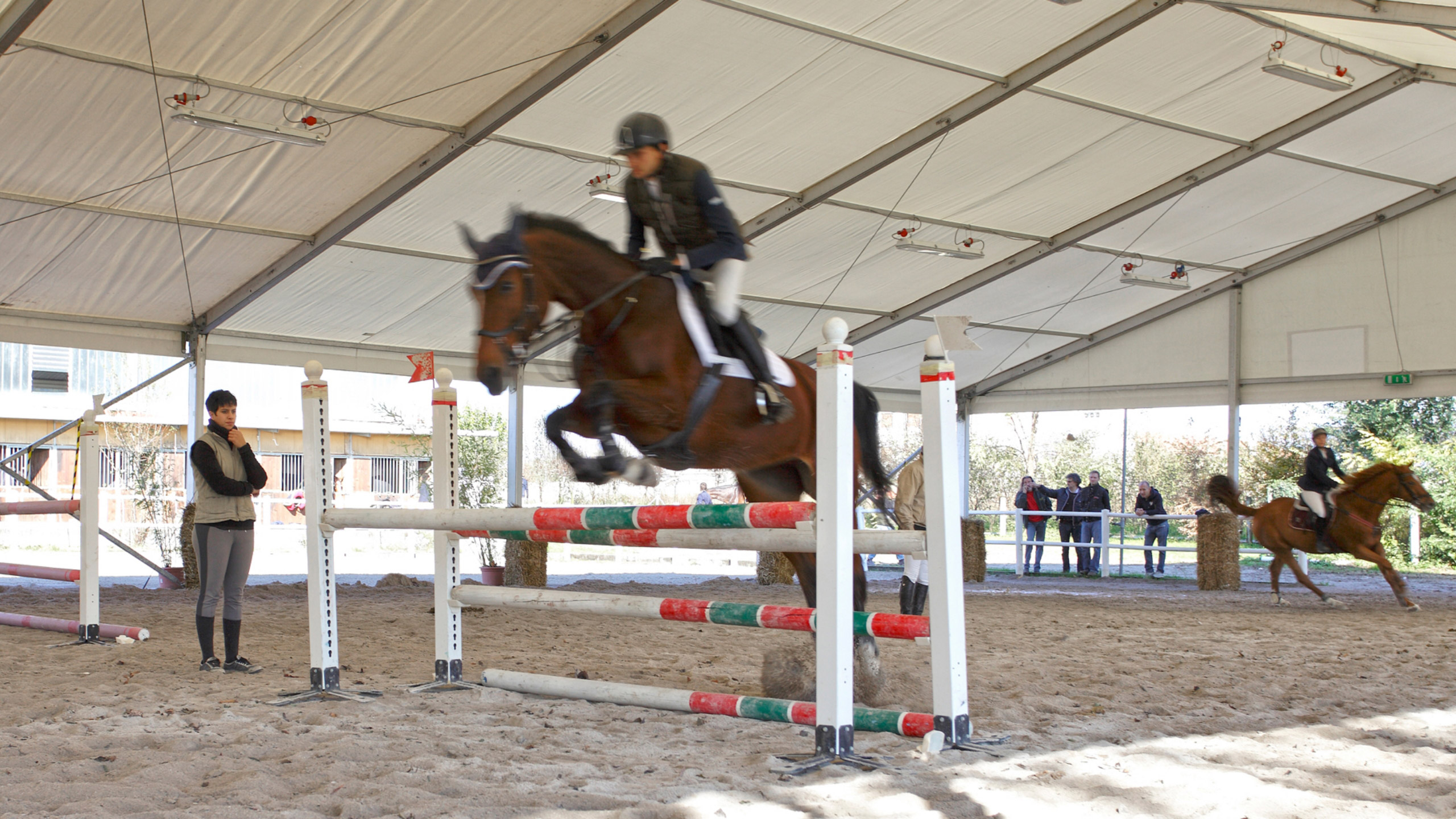 Una persona a cavallo salta un ostacolo in un maneggio coperto, mentre sullo sfondo si vede un altro cavaliere a cavallo e degli spettatori osservano da dietro una barriera.