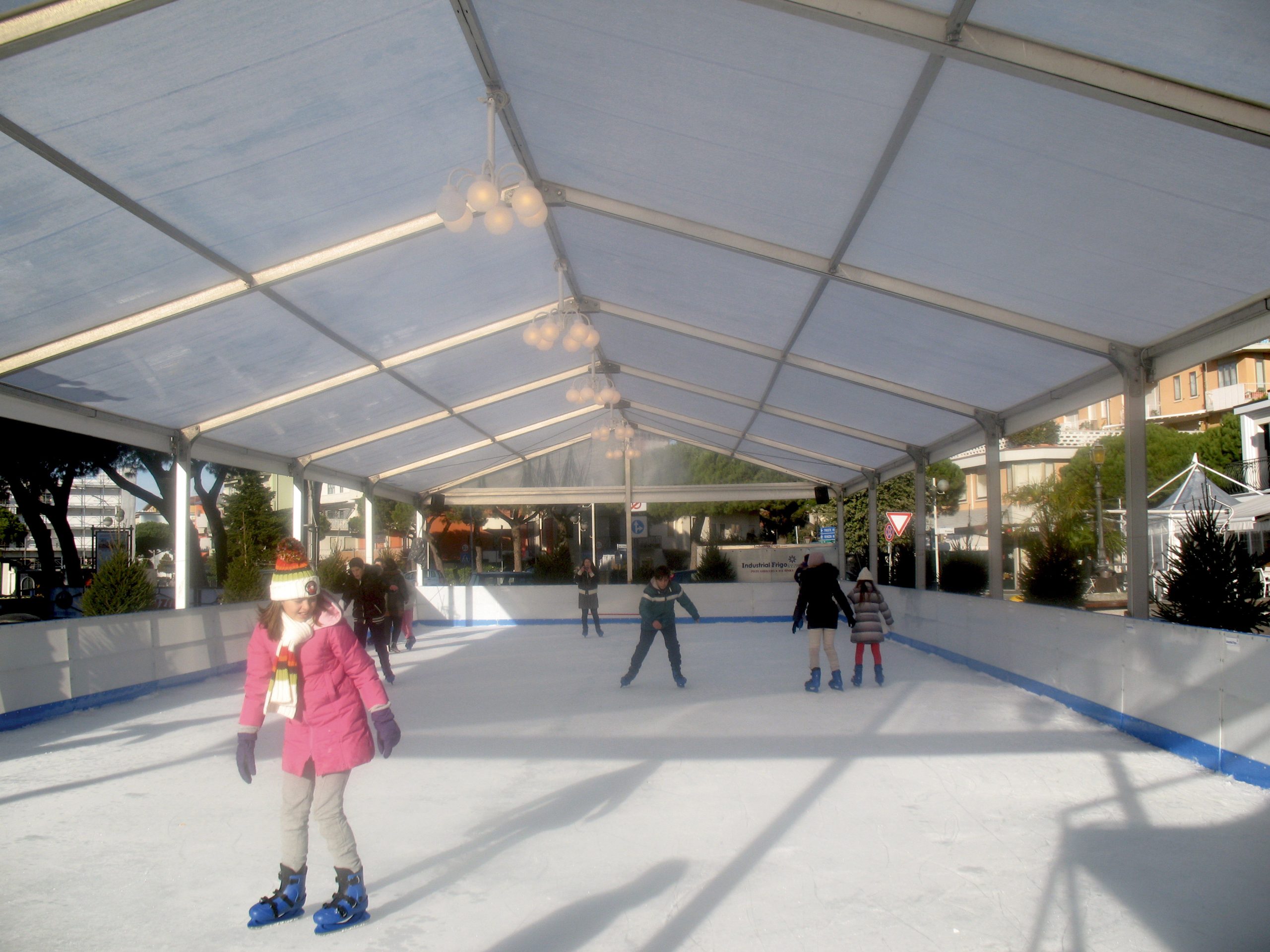 Bambini e adulti pattinano sul ghiaccio sotto una grande tenda con tetto trasparente in una giornata di sole. La pista è circondata da alberi ed edifici, e i pattinatori indossano abiti invernali e pattini blu.