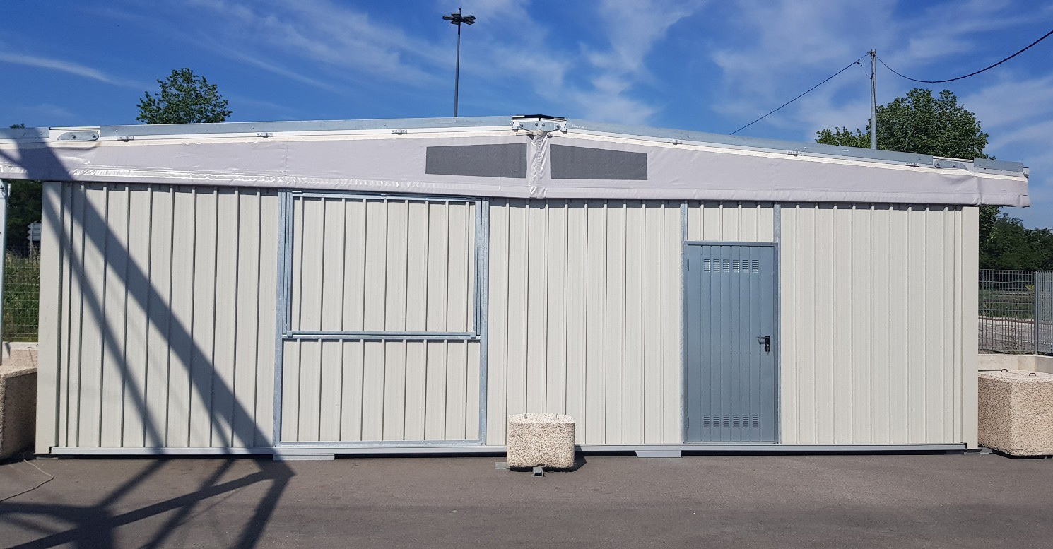 Un edificio prefabbricato in metallo grigio chiaro, con una porta scorrevole sulla sinistra e una porta più piccola, chiusa e dotata di prese d'aria sulla destra, situato su un'area pavimentata sotto un cielo azzurro.