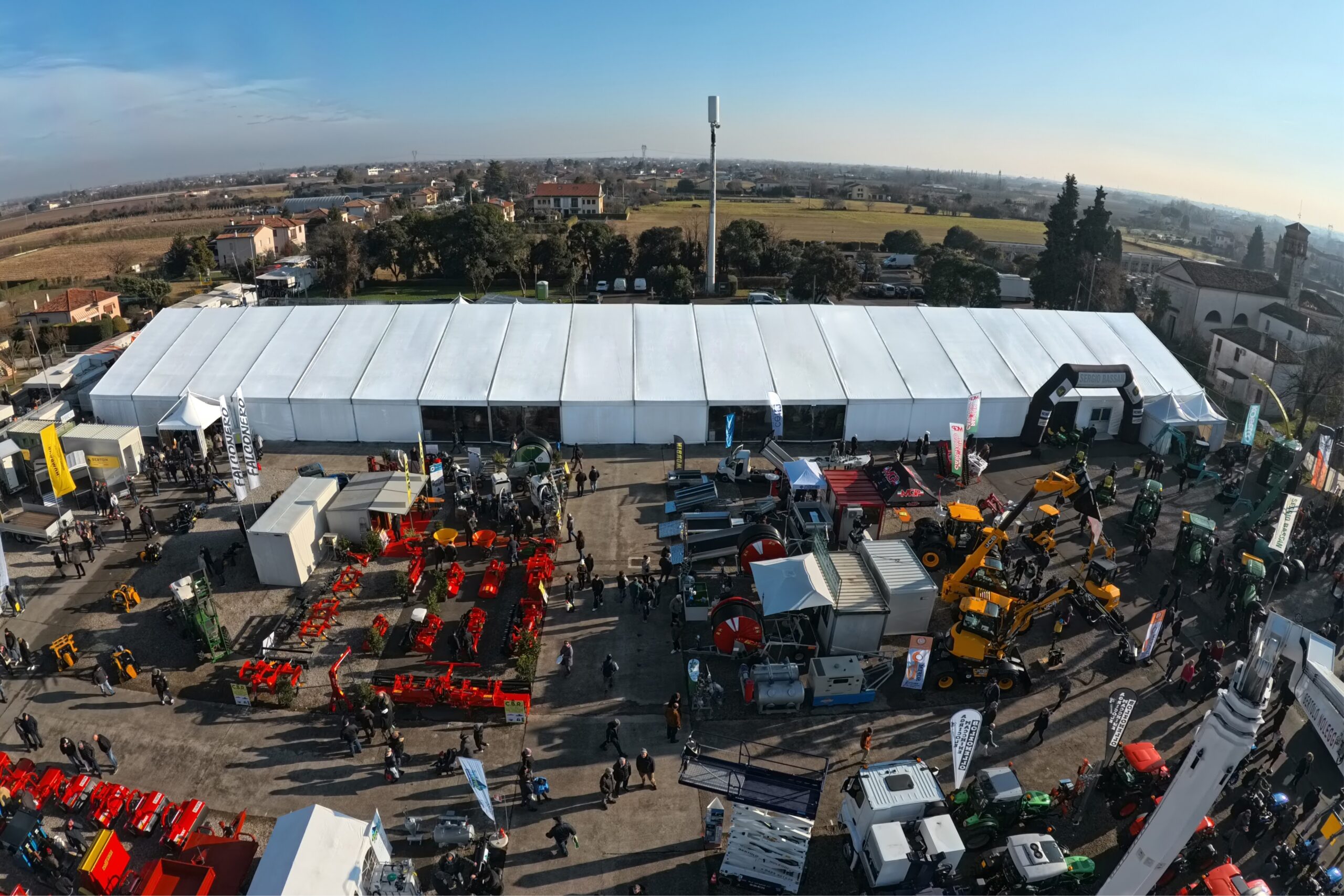 Veduta aerea di una grande esposizione all'aperto con tendoni bianchi e vari macchinari agricoli in mostra. Le persone camminano tra le attrezzature e sullo sfondo sono visibili campi ed edifici sotto un cielo limpido.