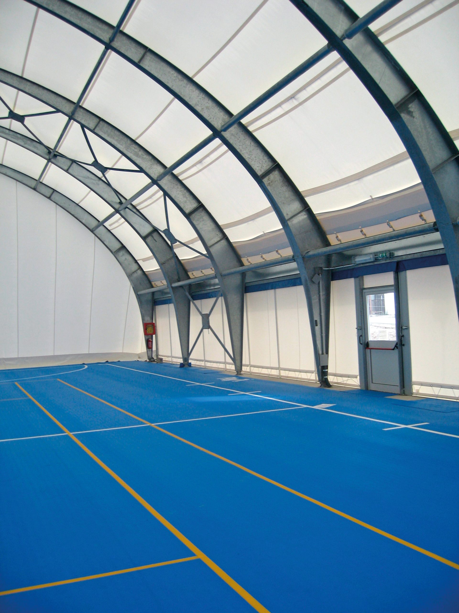Campo sportivo coperto con pavimento blu e linee gialle, coperto da una grande struttura ad arco in metallo e tessuto. Una porta e un estintore sono visibili lungo la parete laterale destra.