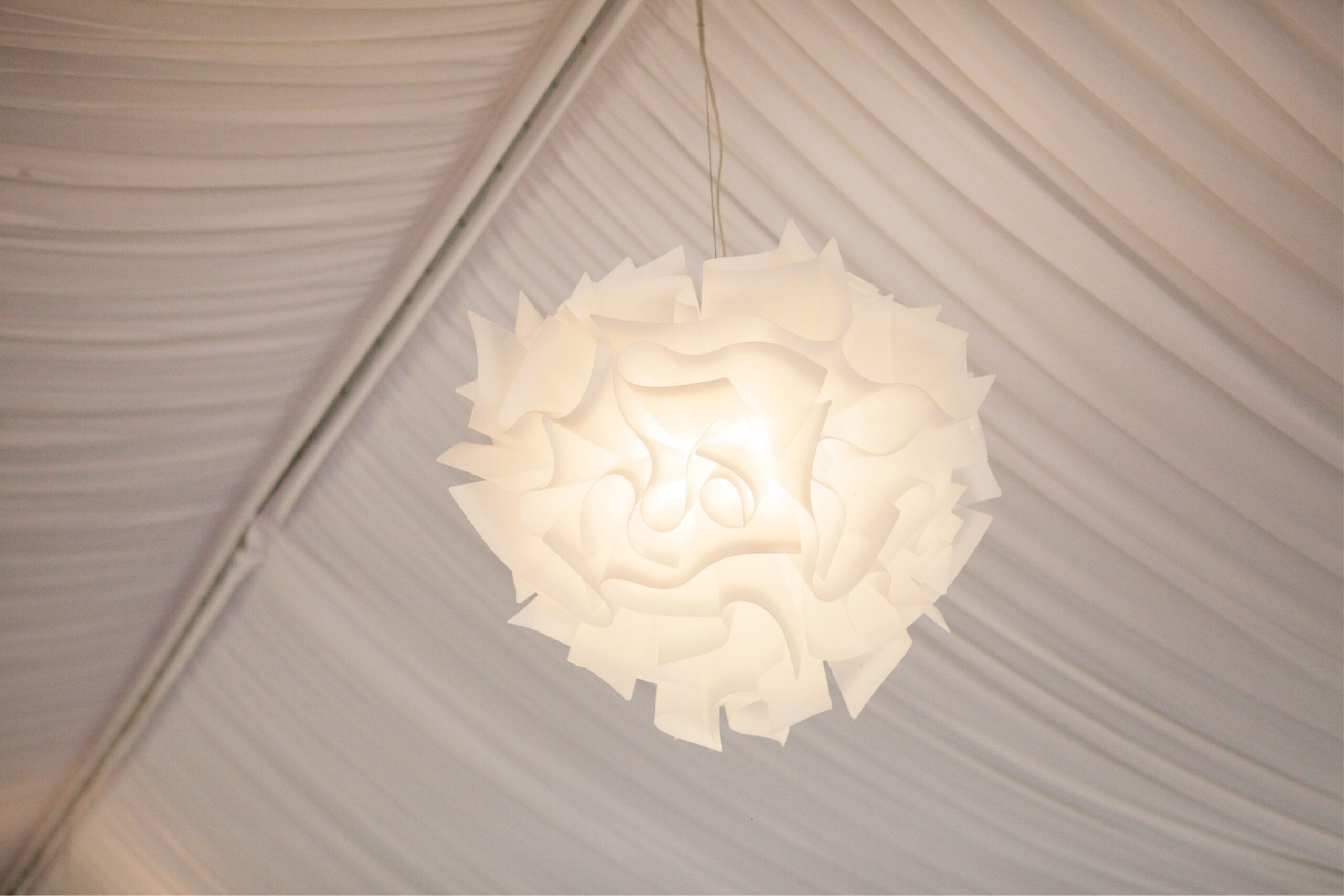 Una moderna e scultorea lampada a sospensione bianca dal design geometrico e ripiegato pende da un soffitto rivestito di tessuto bianco drappeggiato.
