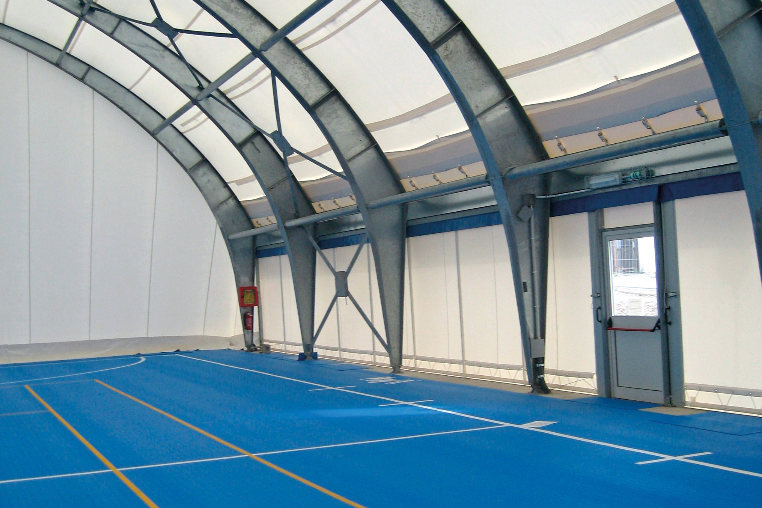 Interno di una struttura sportiva al coperto con pavimento blu contrassegnato per vari giochi da campo, supporti in acciaio ad arco, pareti in tessuto bianco, un estintore e una porta che lascia entrare la luce del giorno.