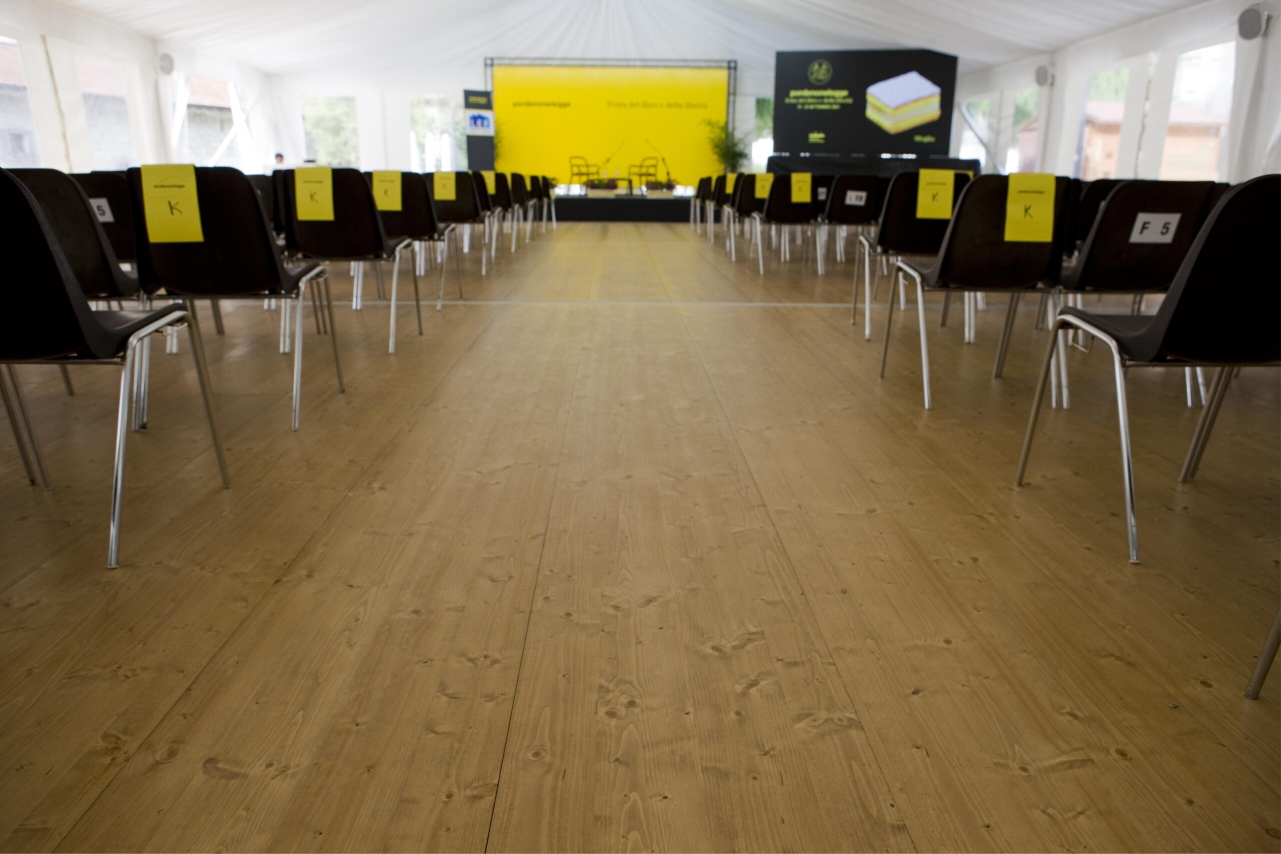 File di sedie vuote con indicatori di posto gialli si affacciano su un palcoscenico all'interno di una tenda per eventi grande e ben illuminata con pavimento in legno. Il palco ha un fondale giallo e schermi con testo e grafica.
