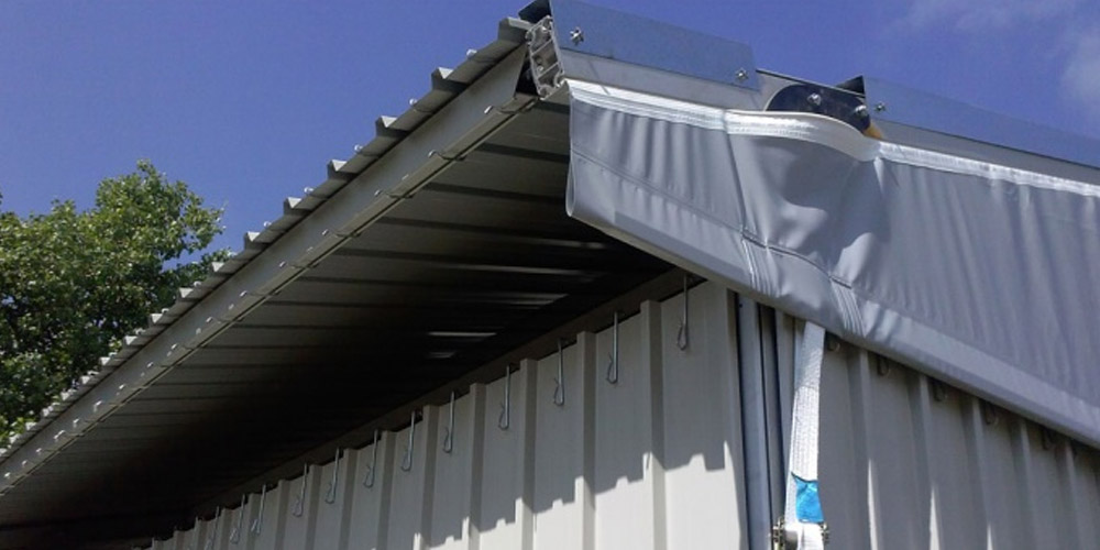 Primo piano di un edificio metallico con tetto ondulato e una copertura in tessuto retrattile grigio parzialmente aperta, fissata vicino al bordo del tetto, sullo sfondo di un cielo azzurro con qualche nuvola e alcuni rami verdi di alberi.